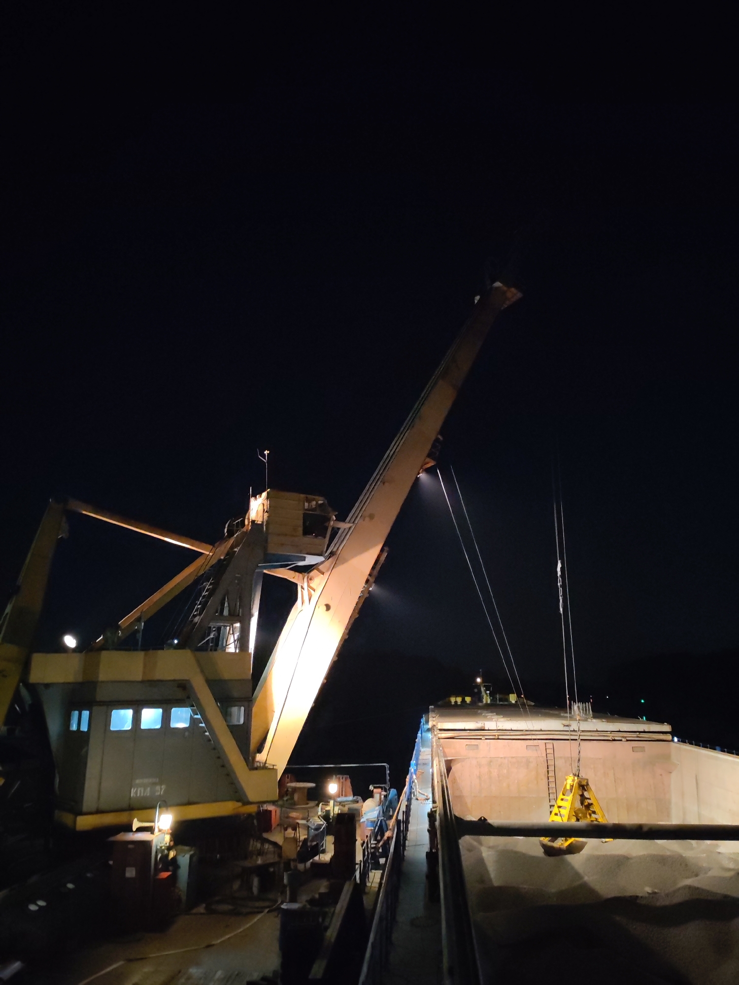 At night in Yakhroma, Russia, a large yellow crane is positioned next to a ship, presumably in the harbor. The crane's arm extends upwards, supporting a bright yellow grab bucket suspended over the ship's hold. The hold appears to be filled with material, possibly sand or gravel. The crane's operational cabin is visible, with lit windows. The scene is illuminated by the crane's work lights and the limited ambient lighting, and a dark background suggests it is late at night. A number 'KPA 57' is printed on the front of the crane.