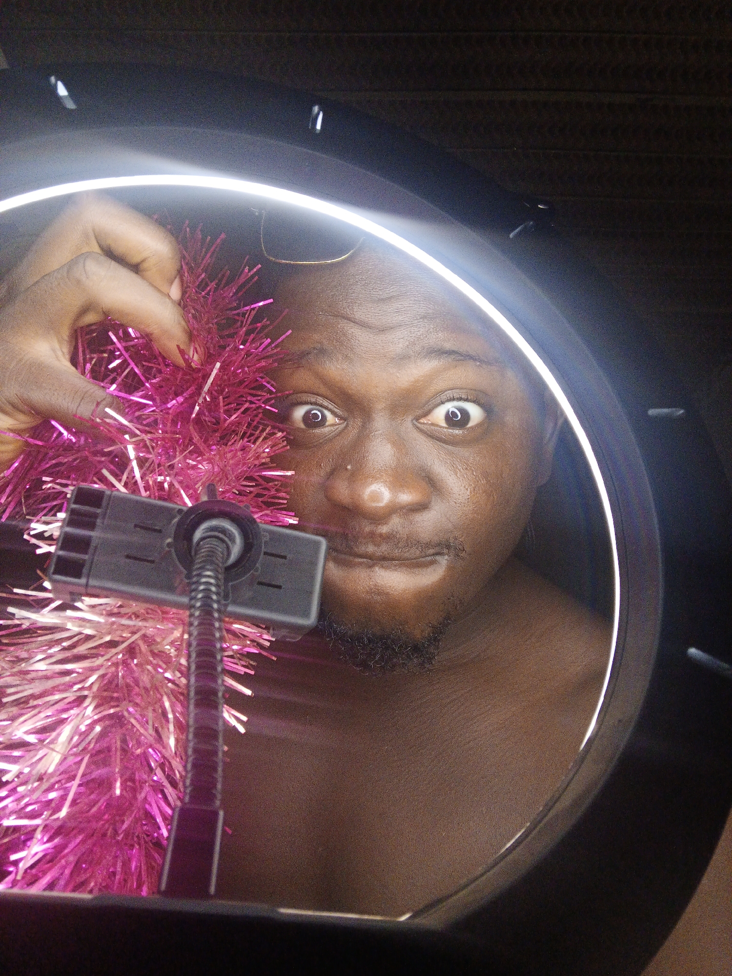 A close-up, low-angle shot captures a dark-skinned person's face from the eyes down, partially obscured by bright pink tinsel and a phone holder, all framed by a large, illuminated ring light against a dark background. The person, who has dark hair and a goatee, is looking directly at the camera with wide, surprised eyes. Their left hand, also dark-skinned, is holding the pink tinsel near their face. The phone holder, black with a flexible gooseneck, is positioned in front of the tinsel. The ring light creates a strong highlight on the person's forehead and nose. The environment is dimly lit except for the ring light.