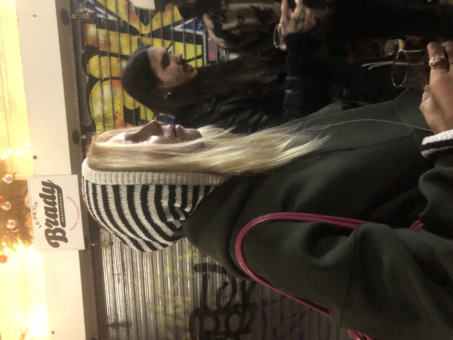 A group of people are situated outside, with one wearing a striped beanie and an olive green jacket with pink straps. They have long blonde hair. Another person, with long dark hair and a black leather jacket, is visible behind. A restaurant sign that reads "Brady" is also present, along with a decorated Christmas tree. The background displays a corrugated metal surface covered with graffiti. A hand adorned with rings is visible on the right edge of the image.