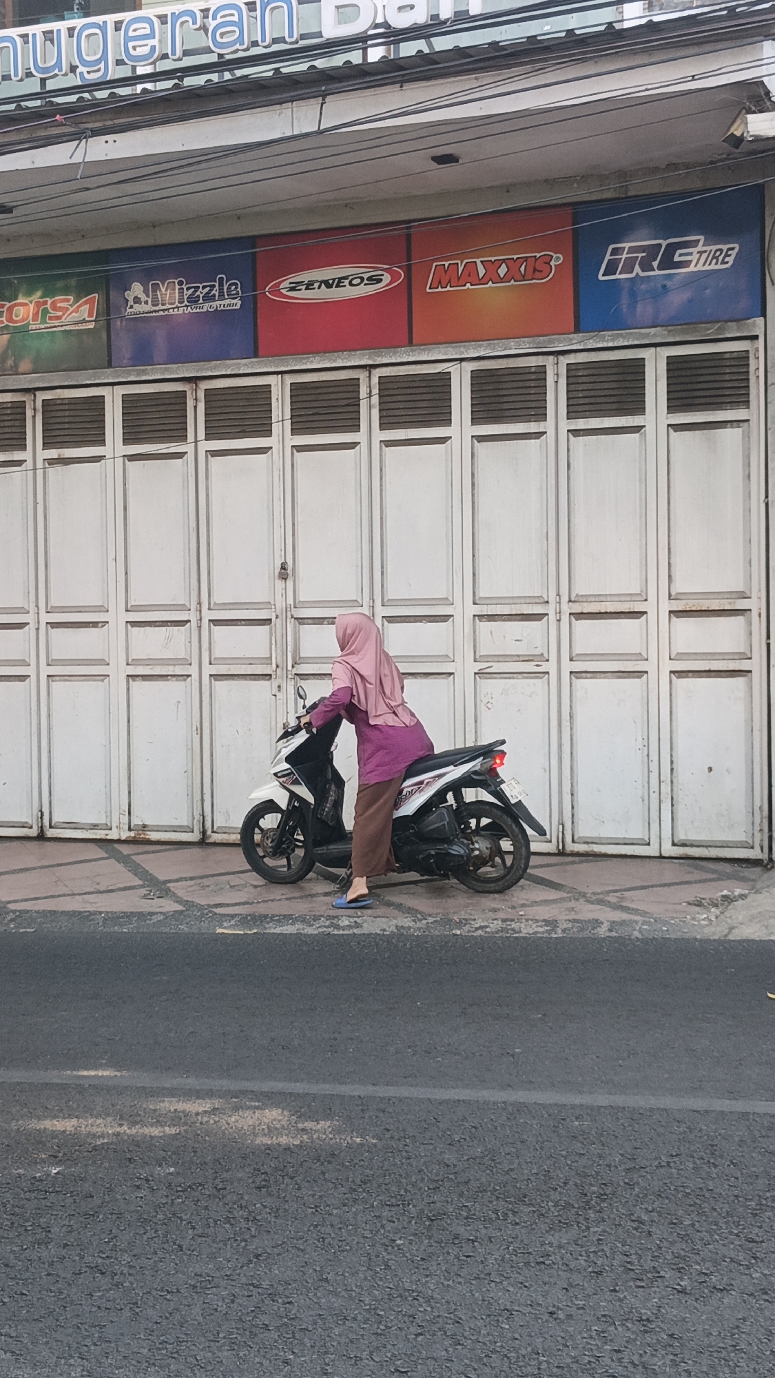 A woman wearing a pink hijab and a purple top, brown pants, and blue flip-flops sits on a white and black scooter parked on the side of the road. Behind her, a row of closed white metal doors extends across the frame. Above the doors, a banner displays logos for "CORSA," "Mizzle," "ZENEOS," "MAXXIS," and "IRC TIRE." The building appears to be a commercial establishment. The road in front of the scooter is asphalt with a white line. The lighting suggests it is daytime.
