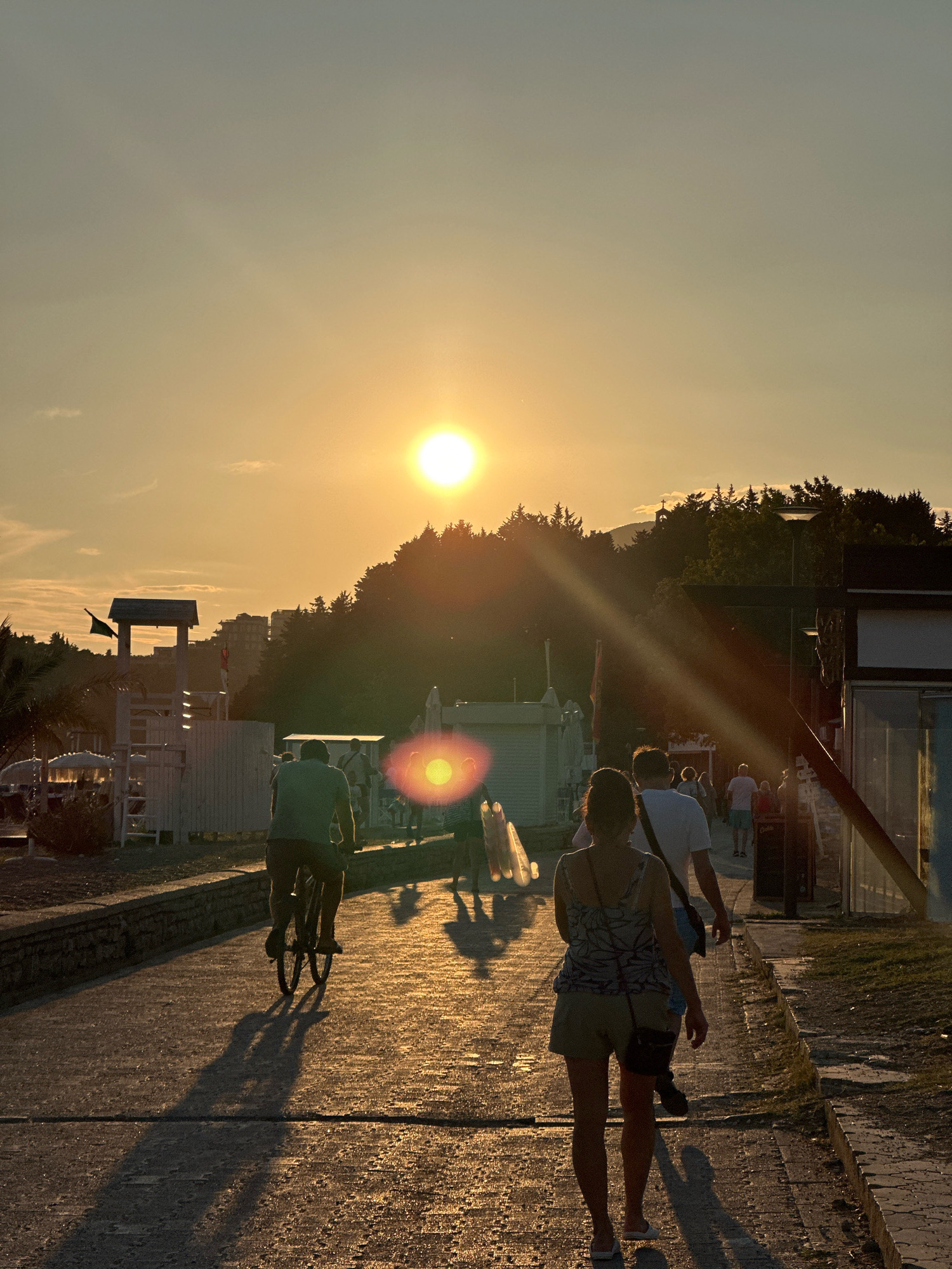 The image depicts a scene at sunset in Boreti, Montenegro. A paved walkway runs through a coastal area, lined with buildings and trees. The sun, low in the sky, casts long shadows and creates a warm, golden light that illuminates the scene.
In the foreground, two women are walking away from the camera on the walkway. The woman on the right is wearing a patterned top and shorts, with a small bag slung over her shoulder. The woman on the left is partially obscured, but appears to be wearing a lighter colored top. Behind them, several other people are walking in the same direction.
Further down the walkway, a person is riding a bicycle towards the camera. This individual is silhouetted against the bright sunlight. In the background, there are several buildings, including what appears to be a lifeguard station with a flag. Lush green trees line the horizon, with some buildings visible behind them. The overall impression is a pleasant evening scene in a tourist area, with people enjoying the sunset.