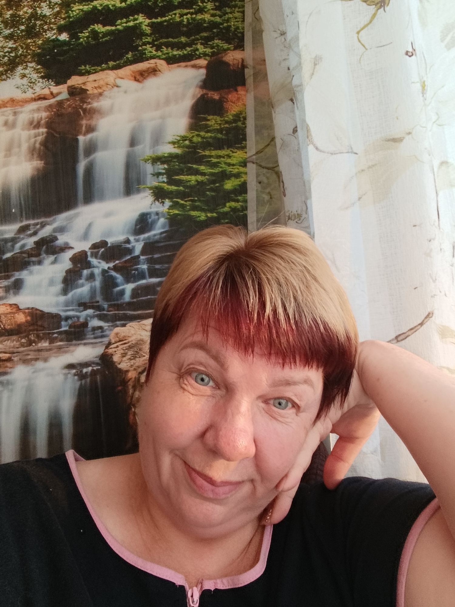 In Svietlahorsk, Belarus, a woman with blonde and red-highlighted hair is taking a selfie. She is wearing a black shirt with a pink trim and zipper. Her right arm is bent, with her hand resting on the side of her head. She is smiling slightly at the camera. Behind her, a wallpaper featuring a waterfall and trees is visible, with a sheer curtain on the right side of the frame.
