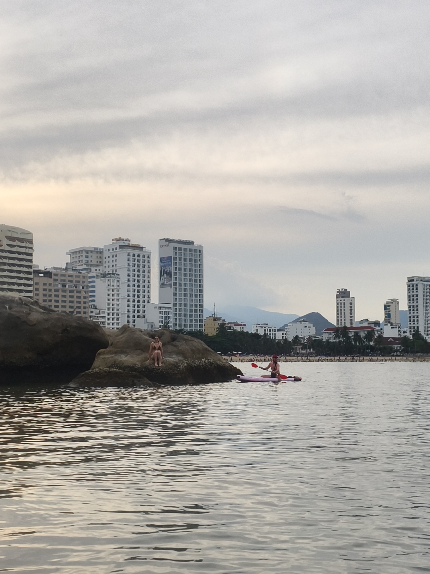 The photograph depicts a coastal scene in Bac Nha Trang Ward, Vietnam. A woman sits on a large rock formation in the water, while a person on a paddleboard is nearby. Several high-rise buildings are visible in the background along the coastline. A beach with many people is present between the water and the buildings. The sky is overcast. The water is choppy and reflects light.