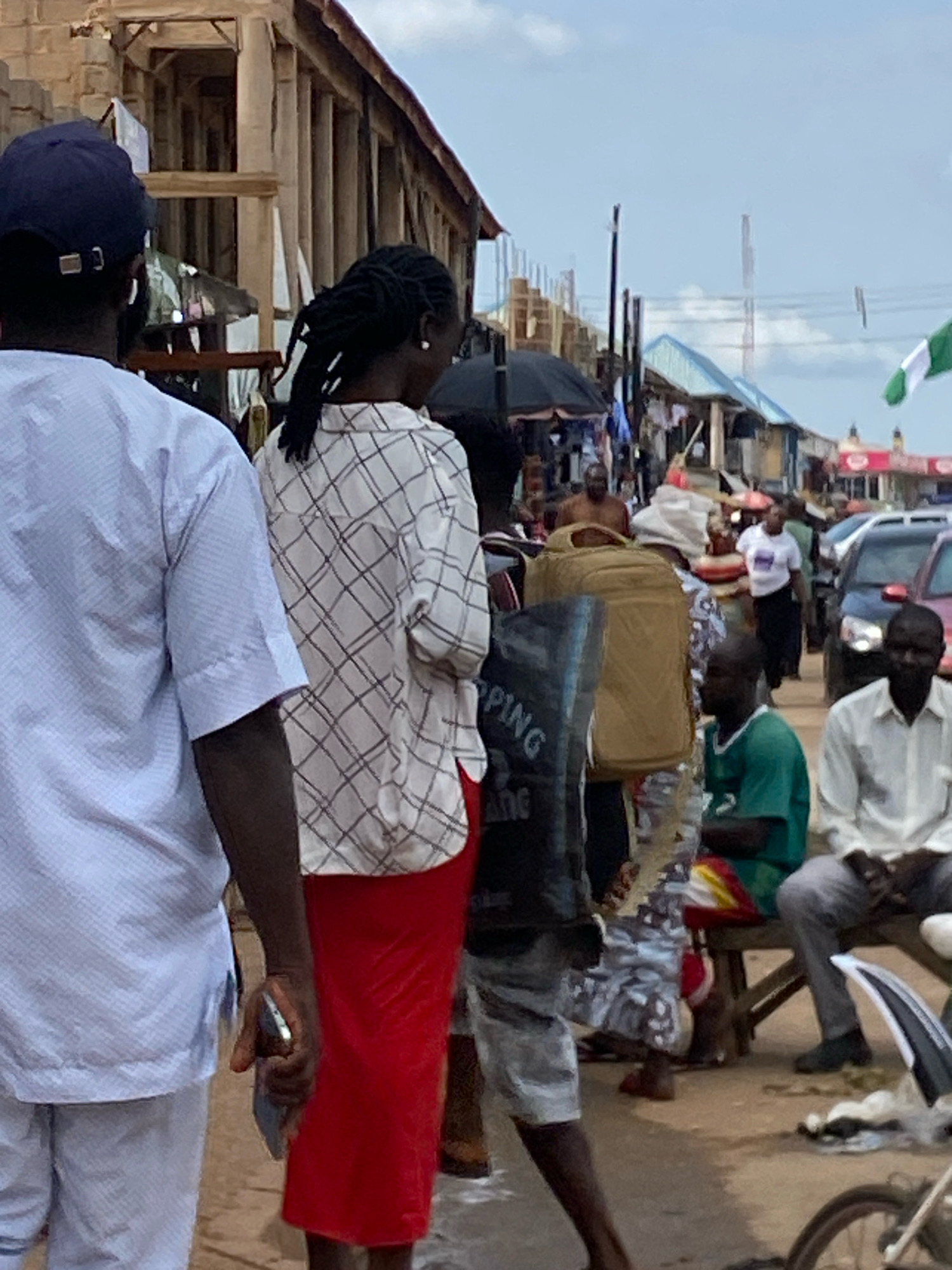 A street market scene is captured in Bukuru, Nigeria, under a partly cloudy sky. In the foreground, a man wearing a white, patterned tunic and pants is viewed from the back and left side. To his right, a woman also viewed from the back and slightly to the side wears a white, checkered blouse and a long red skirt. She is holding a phone in her right hand.
Behind them, a busy marketplace unfolds. Various individuals are visible among stalls and parked vehicles. A man in a green shirt is seated on a bench to the right, facing left. Next to him, a man in a white shirt and grey pants is also seated. Several vehicles, including cars and a motorcycle, are present on the street. Market stalls with various goods are set up, and umbrellas are open, suggesting protection from the sun. A Nigerian flag, green and white, is visible in the background, attached to a pole. The architecture appears to be a mix of semi-permanent structures and open-air stalls, typical of a Nigerian market. The overall atmosphere is that of a lively and active commercial hub.