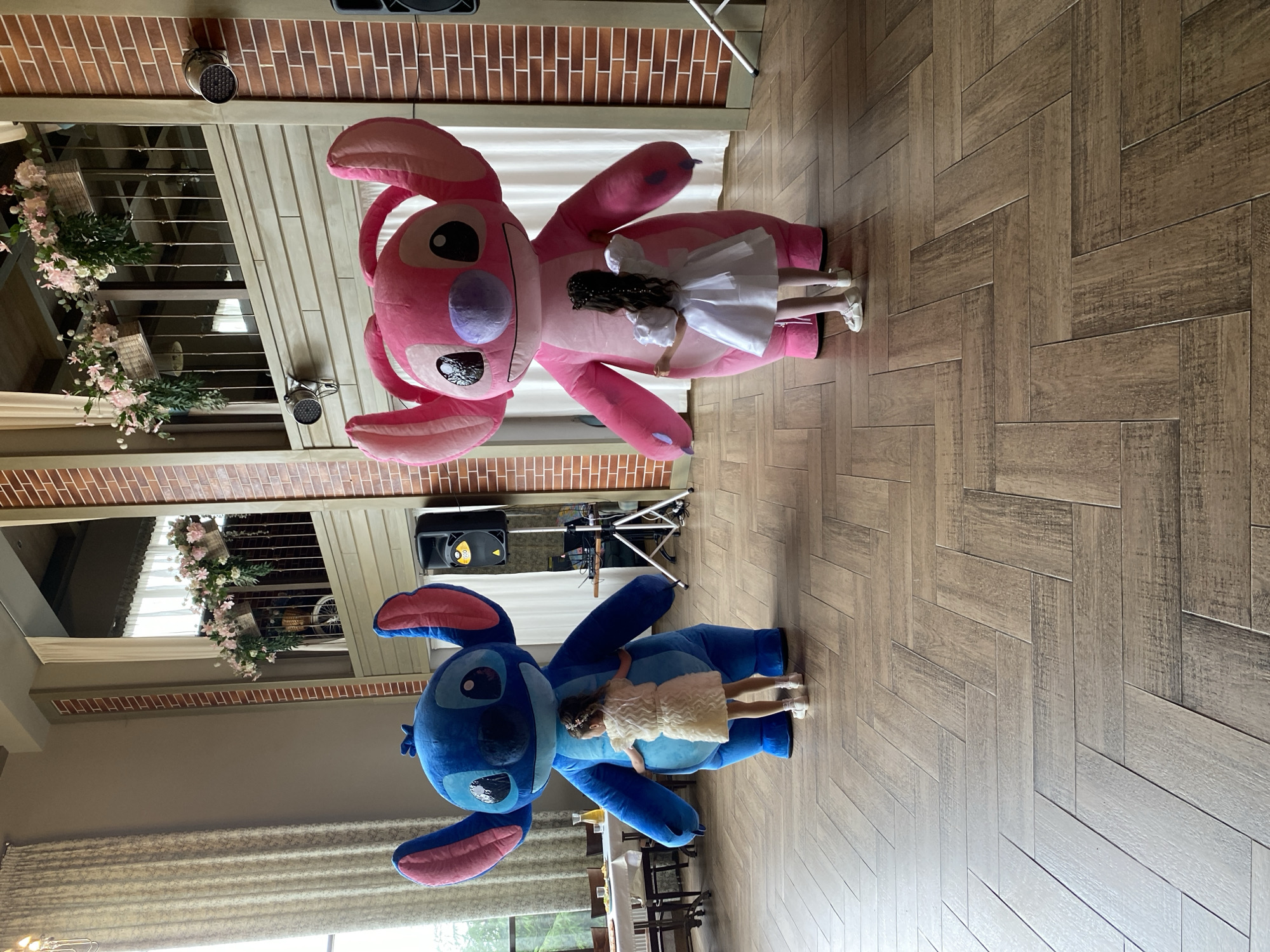 Two children are lying on large stuffed animal versions of the characters Stitch (blue) and Angel (pink) in what appears to be a venue decorated for an event. The floor is a light brown herringbone pattern. In the background are brick walls, large windows, and floral decorations. A speaker and sound equipment are also visible. Based on available information, the location is possibly Votkinsk, Russia.