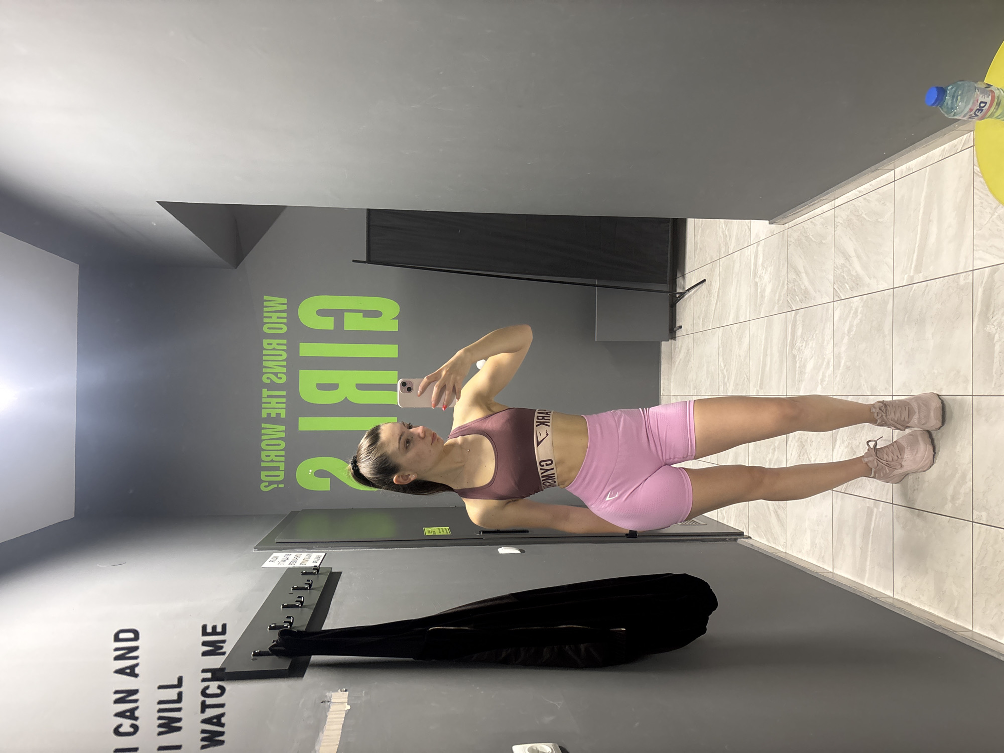 A woman in workout attire takes a selfie in a gym. The gym's decor features gray walls with motivational text in neon green and white. A water bottle sits on a yellow table or surface in the background. A black bag is hanging on the wall. The gym's exact location in Varna, Bulgaria, cannot be determined from the image.