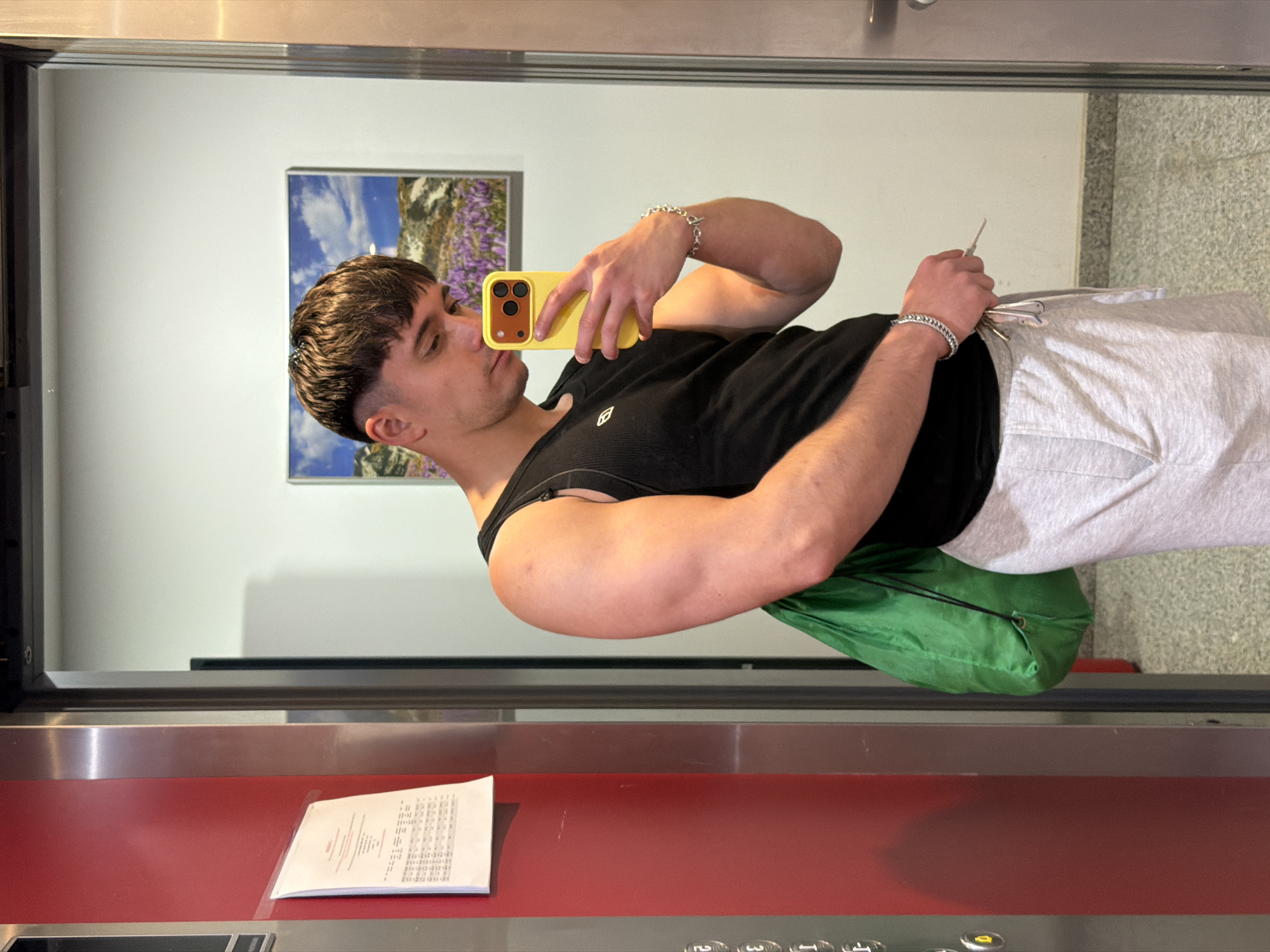 A young man stands in front of a mirror in an elevator. He is holding a yellow cell phone up to his face, presumably taking a selfie. He is wearing a black tank top and light gray sweatpants. He is holding a set of keys in his left hand. There is a green bag beneath his left arm. The elevator has a red control panel with buttons. There is a piece of paper on the control panel. The wall behind the man and elevator is white with a landscape painting hanging on it. The floor appears to be a terrazzo material. The location is unknown, and there is no evidence in the image to suggest it is Sofia, Bulgaria.
