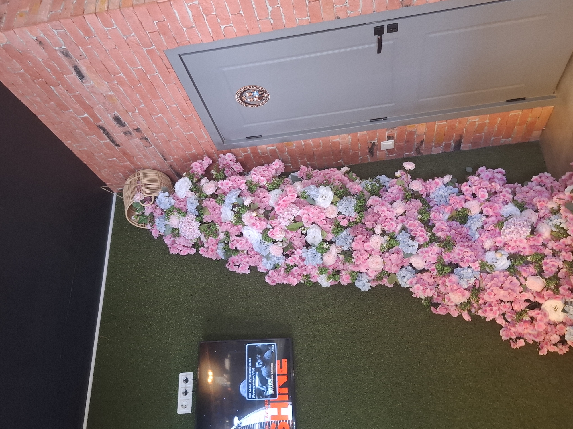 In a dimly lit interior space, a wall covered in artificial green turf is visible on the left. A black flat-screen television is mounted on this wall, displaying an advertisement with text in Russian. To the right of the turf wall, a tall, vertical arrangement of artificial pink and light blue flowers cascades down from a small wicker basket. This floral display is positioned next to a reddish-orange brick wall. A light gray door with black hardware is set into the brick wall, featuring a small oval plaque with two figures on it. A light switch is visible on the brick wall near the bottom of the door. The ceiling is black. The scene is likely indoors, possibly in a commercial establishment.