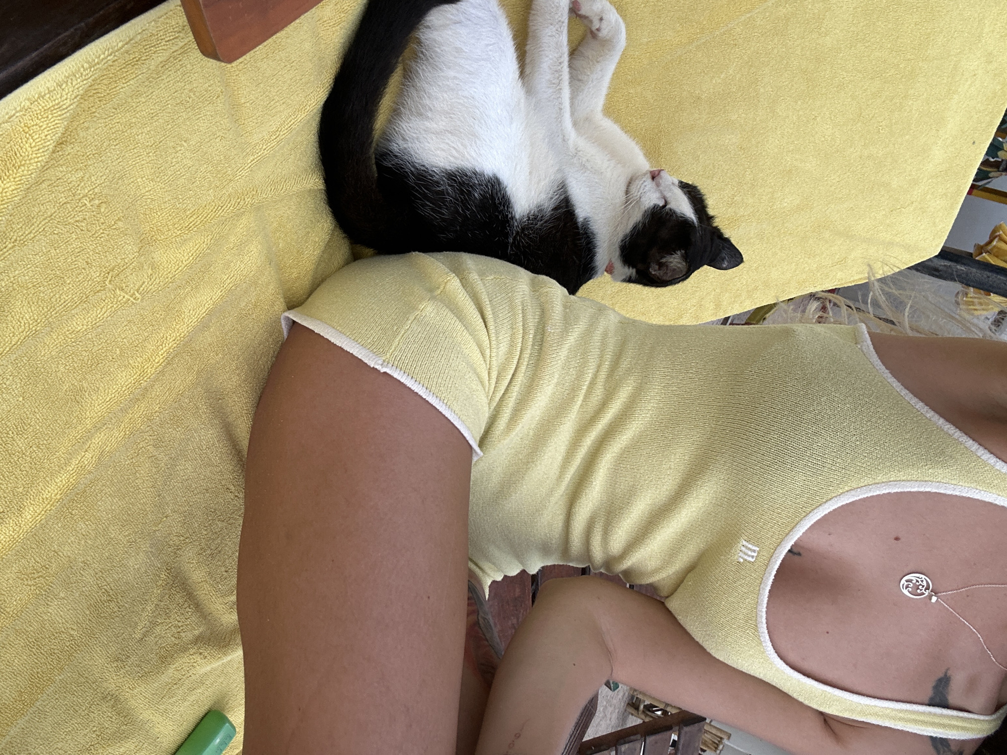 A black and white cat naps on a yellow towel next to a woman wearing a yellow outfit. The woman has a necklace with a circular pendant and a tattoo on her chest and arm. The cat is lying on its side with its eyes closed. The background is blurred but suggests an outdoor setting with some furniture.