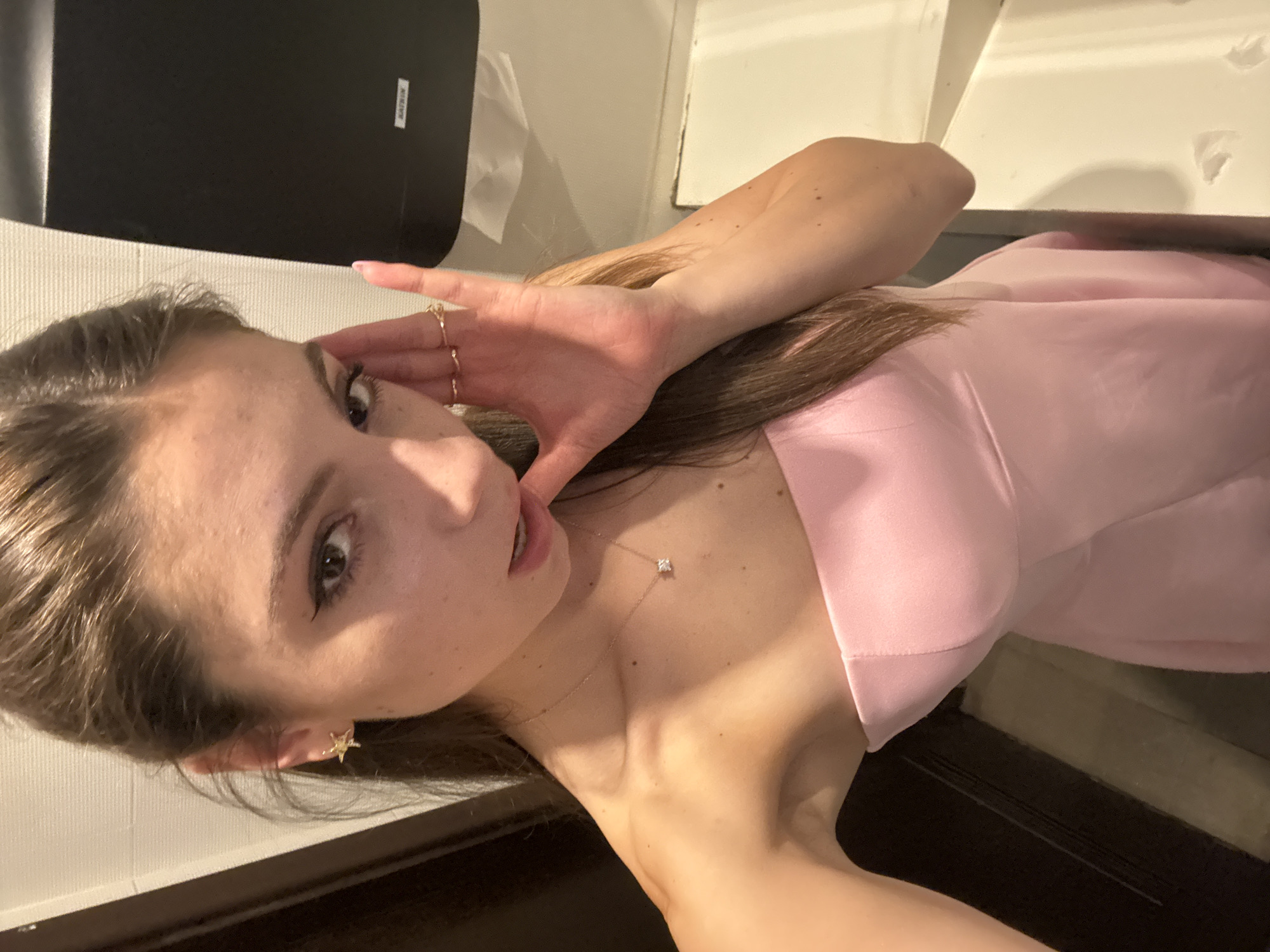 A young woman is posing for a selfie in a bathroom. She is wearing a light pink strapless dress, a delicate gold necklace with a square pendant, a gold star earring, and multiple gold rings on one hand. Her hand is raised to the side of her face, partially obscuring it. The bathroom features a black paper towel dispenser on a white wall. The scene is not identifiable as being in Shumen, Bulgaria.