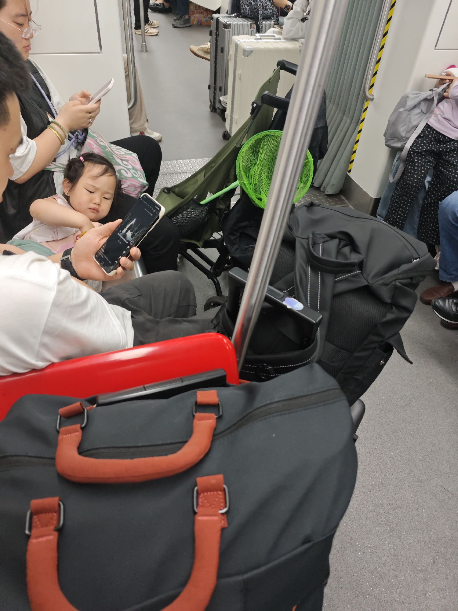 On what appears to be public transportation, a young child sits on an adult's lap, looking at a smartphone held by the adult. Another adult sits next to them. The setting includes luggage, bags, a stroller, and standing passengers. The location is Unknown, Unknown.