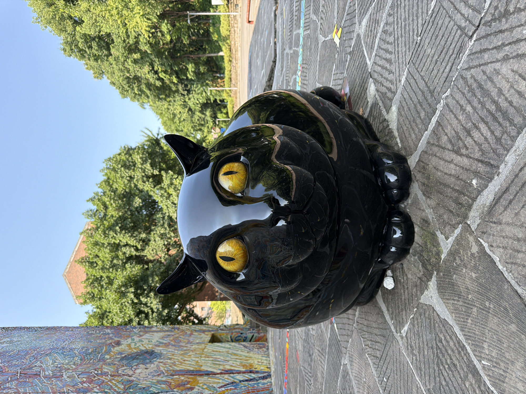 A glossy black cat sculpture with large yellow eyes sits on a paved courtyard. The sculpture is positioned in the center of the frame, facing the viewer directly. The courtyard is paved with large, grey stones laid in a herringbone pattern. In the background to the left, a colorful mosaic mural adorns the side of a building. Further back, green trees and a brick building are visible under a clear blue sky. To the right of the sculpture, more trees line a path with a road and lampposts in the distance. The scene appears to be an outdoor public space. The location is the Chongqing High-tech Zone, China.