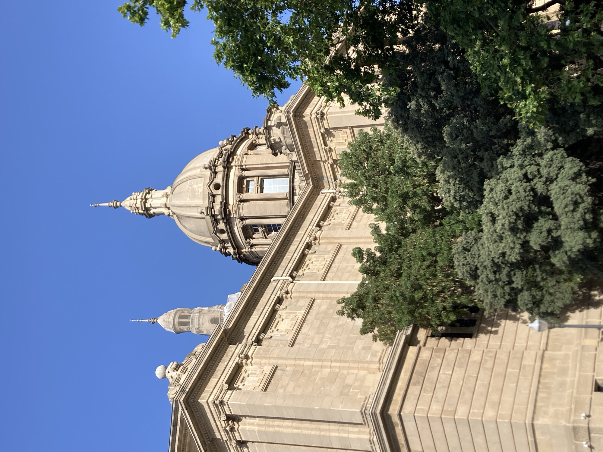 The image captures a low-angle view of the National Art Museum of Catalonia (MNAC) in Barcelona, Spain, under a clear blue sky. The grand building, constructed of light-colored stone, features classical architectural elements, including columns, cornices, and decorative carvings. A prominent dome with a spire dominates the upper right portion of the building, while another, narrower tower with a similar spire is visible behind it on the left. Lush green trees partially obscure the lower sections of the building, with their foliage creating a contrast with the stone structure. The lighting suggests it is daytime, with the sun casting subtle shadows. No people are visible in the frame.