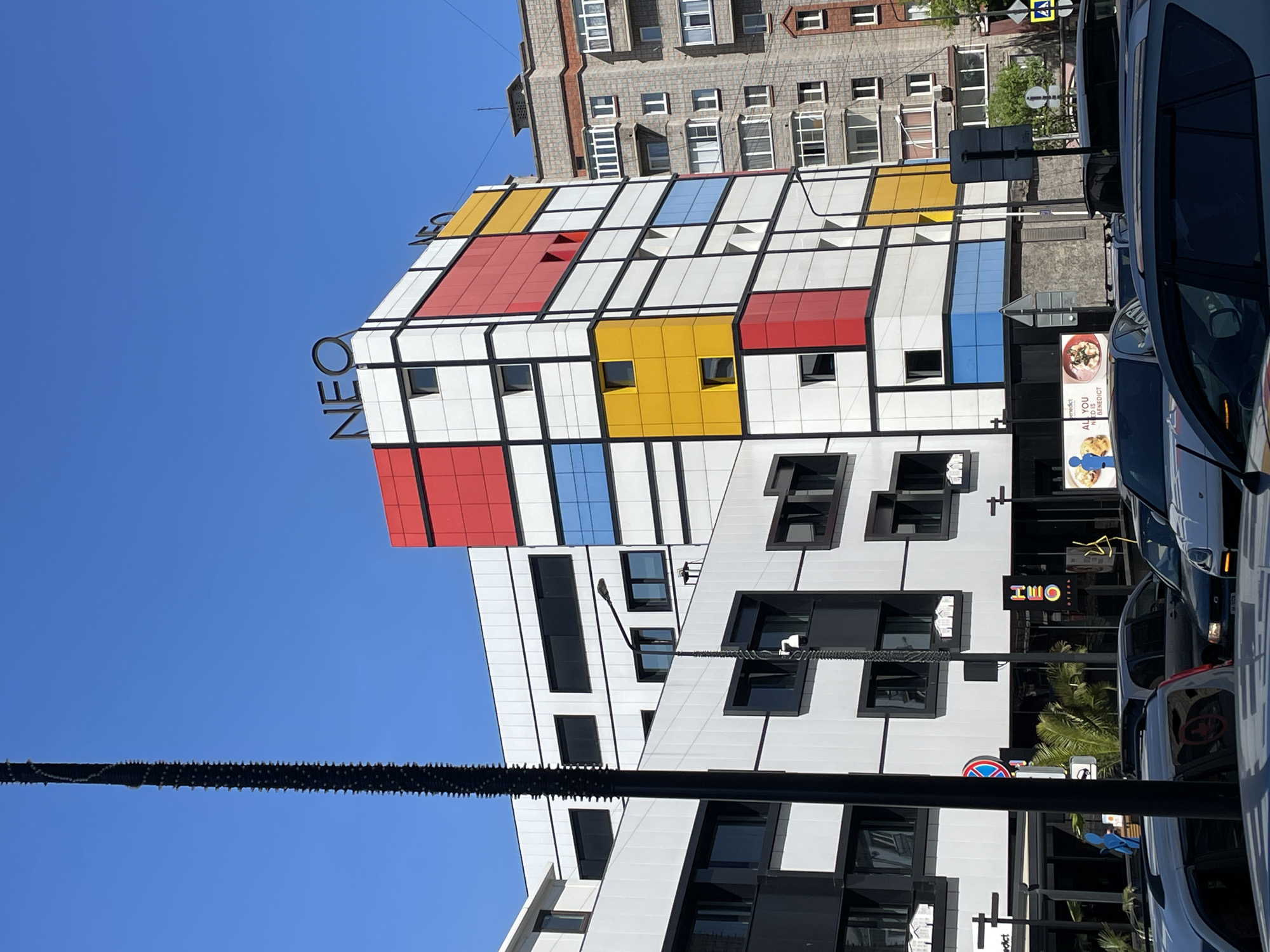 This image captures a vibrant urban street scene, centered around a striking modern building. The time of day is clear and sunny, indicated by the bright blue sky and crisp shadows.
The main subject is a multi-story building featuring a facade inspired by the De Stijl art movement, characterized by a grid of black lines framing rectangles of primary colors – red, yellow, and blue – interspersed with white and dark-framed windows. The word "NEO" is prominently displayed in large, dark letters on the roofline of this building. Behind it, an older, more traditional building with a textured, possibly brick facade and numerous smaller windows stands.
In the foreground and to the right, parts of several vehicles, including what appears to be a white or light-colored van and a dark car, are visible, suggesting a busy street or parking area. A thick, dark cable, possibly adorned with small, spike-like elements, stretches horizontally across the middle ground against the clear sky.
The lower levels of the modern building are predominantly white with large, dark rectangular windows. Visible street furniture includes a black lamppost or utility pole, a yellow square traffic sign, and a circular red and blue street sign. Green foliage, possibly from a palm tree, can be seen near the base of the buildings.
Visible text elements include "NEO" on the modern building's roof and near its entrance, and "H₂O" on a smaller sign. Through a car window, a poster advertising "Benedict" and stating "ALL YOU NEED IS BENEDICT" is discernible. No specific activity or event is taking place beyond the static portrayal of a city street in Krasnoyarsk, Russia, under good weather conditions.