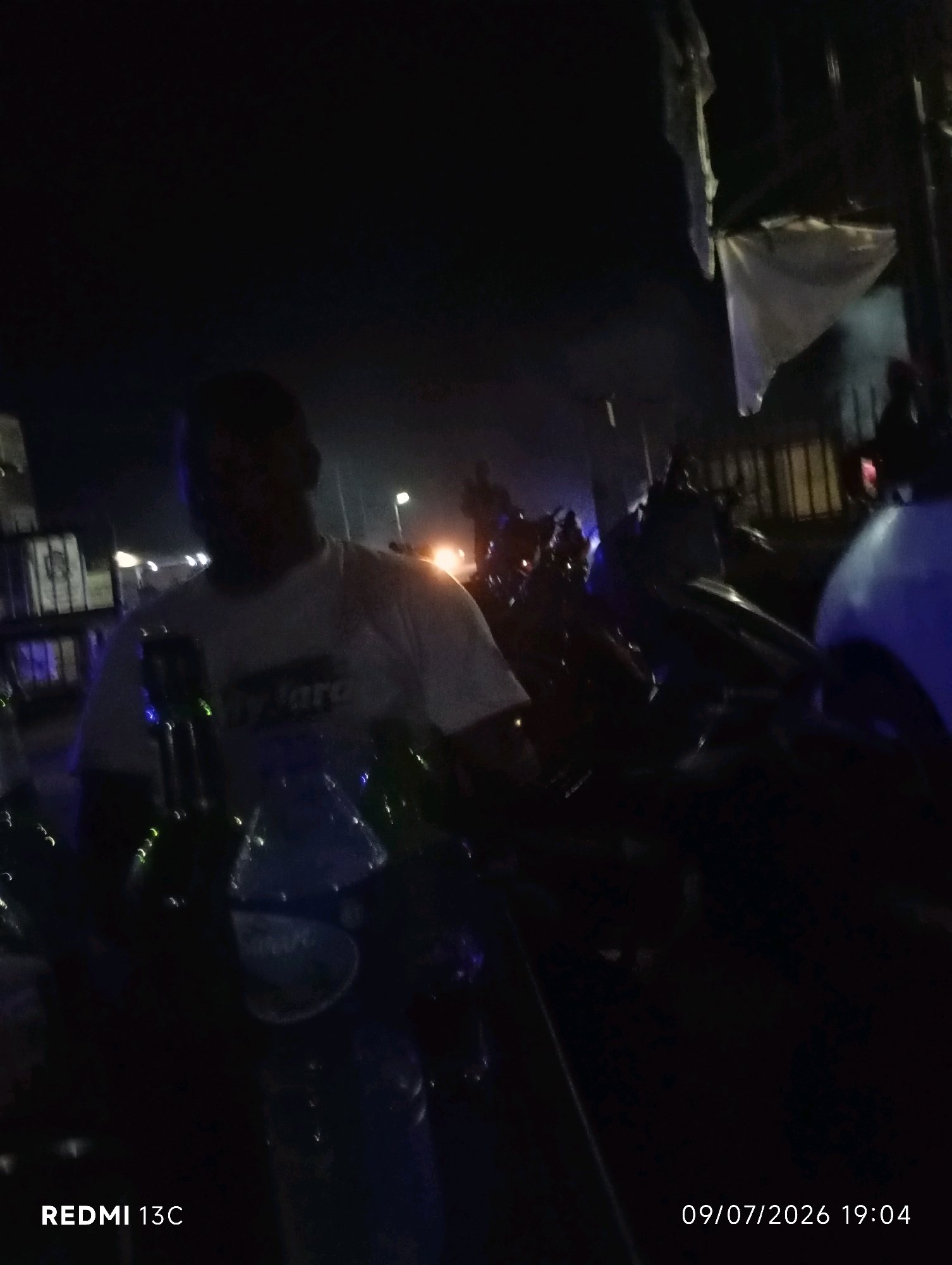 In the city of Shendam, Nigeria, at 7:04 PM on July 9, 2026, this image captures a dimly lit outdoor scene. A person, silhouetted against a dark background, stands in the foreground, wearing a light-colored t-shirt with text that is partially obscured. In front of this individual, several green glass bottles are visible, some appearing to be partially filled. The environment suggests a public space, possibly a street or an area with some form of temporary shelter, indicated by the presence of structures and a tarp-like material in the background. The overall atmosphere is dark, with artificial lights providing illumination in various areas. The image also includes the text "REDMI 13C" in the lower left corner, indicating the device used to take the photograph.