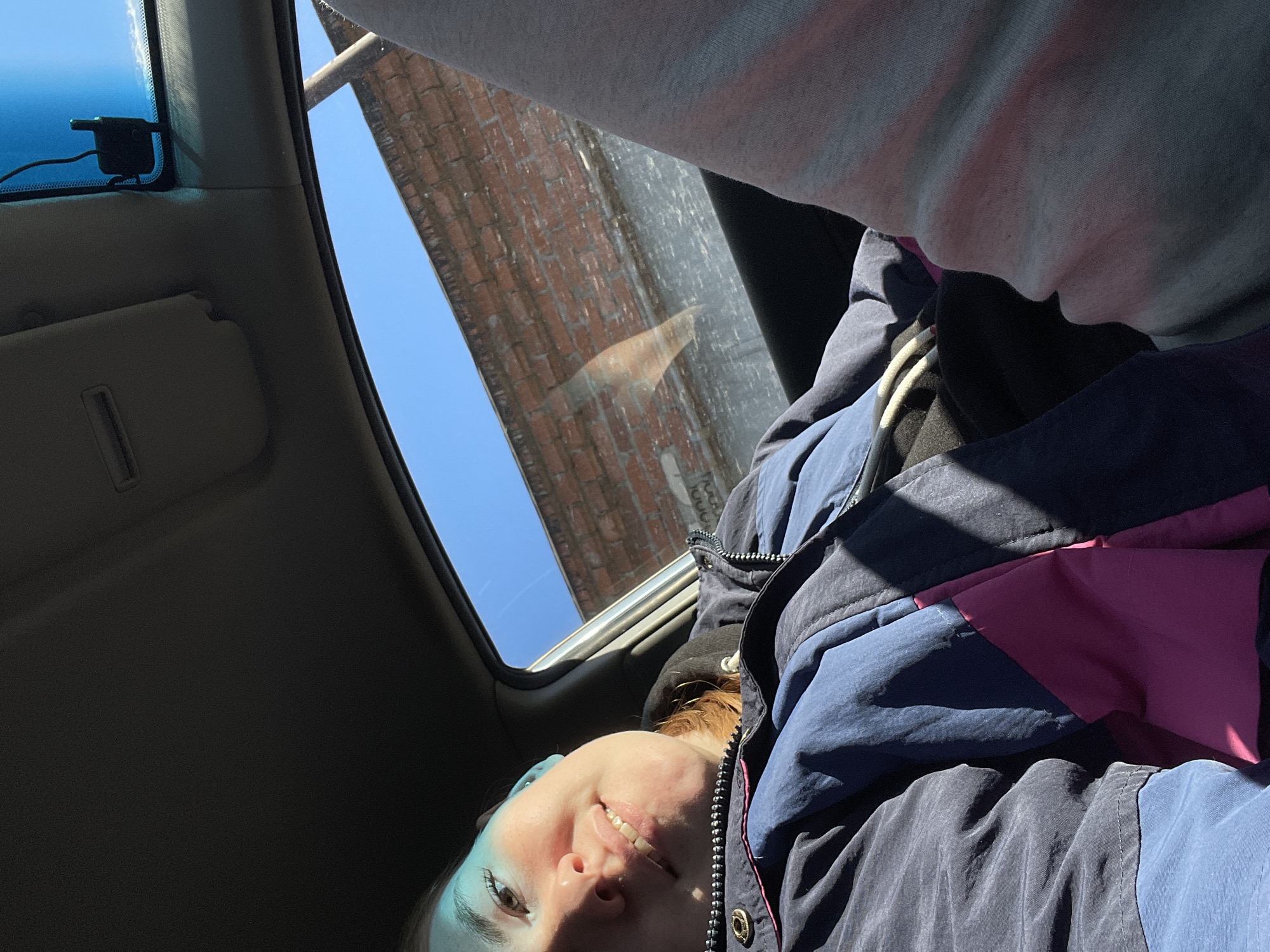 A woman is taking a selfie while sitting in a car with the sunroof open. The sunroof reveals a brick building and blue sky above, possibly in Chaikovsky, Russia. The woman is wearing a blue and pink jacket. A rearview mirror and sun visor are visible on the car's ceiling. Another person is partially visible in a gray shirt above the woman.