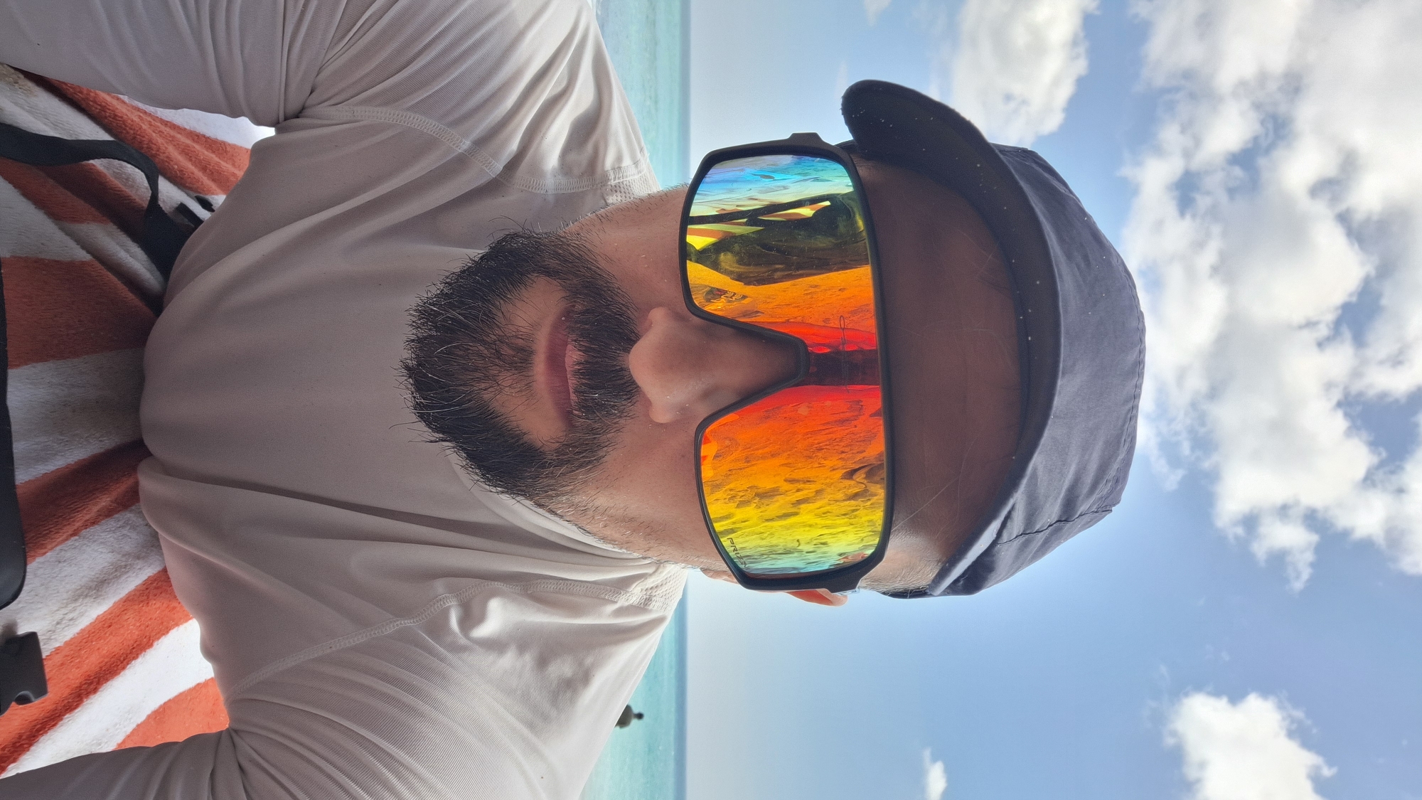 A man wearing a dark baseball cap, large mirrored sunglasses with a rainbow reflection, and a long-sleeved white shirt is taking a selfie. He has a beard and mustache. He is positioned in front of a beach scene with turquoise water and a cloudy sky. The man appears to be lying down on a towel with orange and white stripes. In the background, a person can be seen standing in the water, and a distant island or landmass is visible on the horizon.