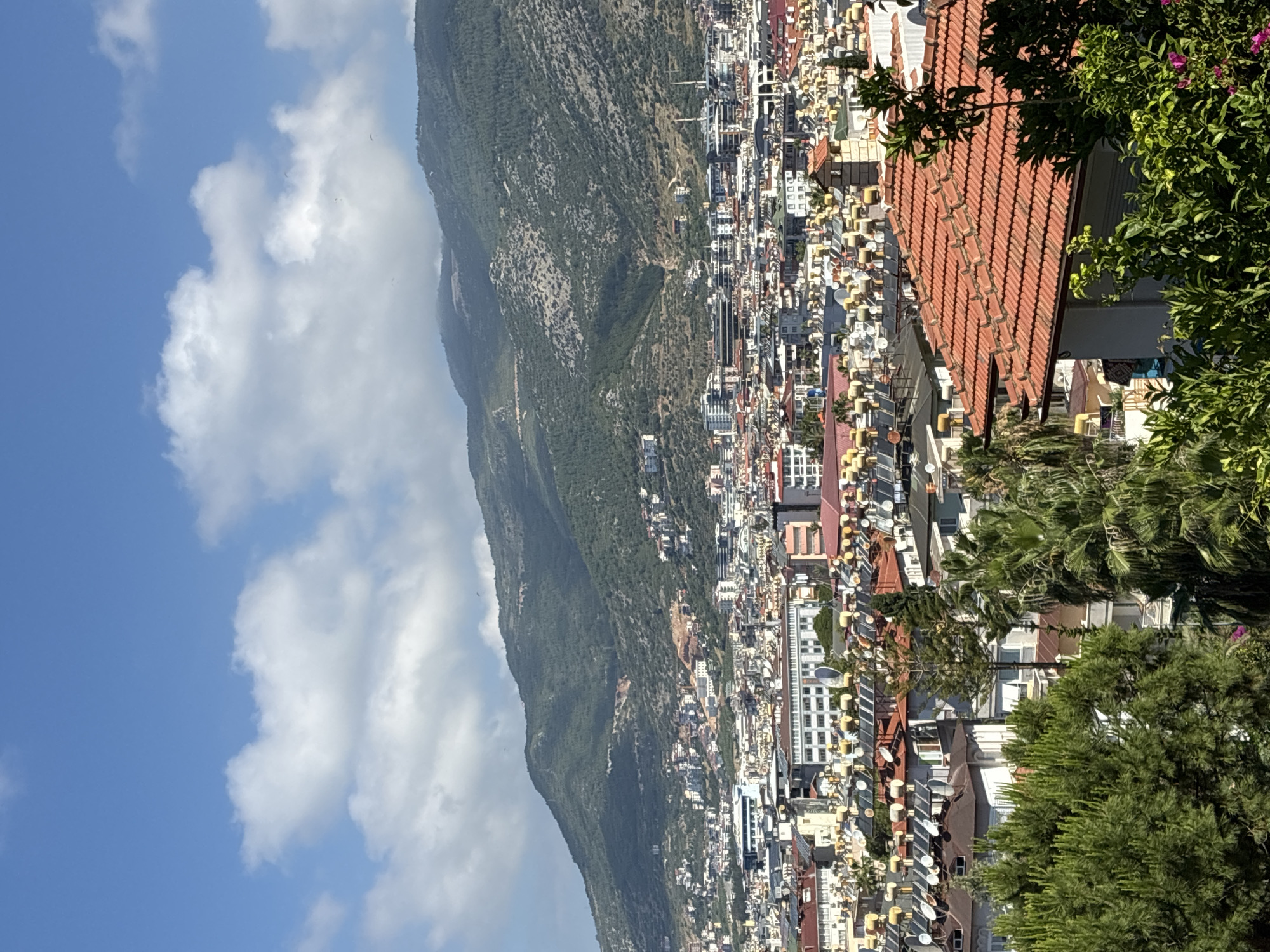 The image presents a panoramic, elevated view of a vibrant coastal city nestled at the base of a majestic, verdant mountain range, under a bright blue sky with scattered white clouds. The perspective looks down over the urban area towards the natural landscape.
The upper portion of the frame is dominated by a clear blue sky, interspersed with fluffy white cumulus clouds. Several small paragliders can be faintly discerned high in the sky against the clouds, indicating recreational activity. Below this, a sprawling, green mountain range extends horizontally across the middle of the scene. Its slopes are covered in dense vegetation, appearing rugged in places.
In the foreground and extending towards the base of the mountains is a densely packed urban area. Numerous low-to-medium-rise buildings with a mix of red-tiled and flat roofs characterize the city's architecture. Many buildings feature balconies, and their rooftops are adorned with numerous satellite dishes and some solar water heaters. Scattered throughout the urban landscape and along the mountain slopes are various trees, including what appear to be palm trees and other dense green foliage, adding to the Mediterranean feel.
The scene is brightly lit by natural sunlight, suggesting a clear day, likely in the morning or early afternoon. The overall atmosphere is bright and energetic, typical of a lively coastal settlement. Given the location context, this view depicts a part of Alanya, Turkey. There is no visible text in the image.