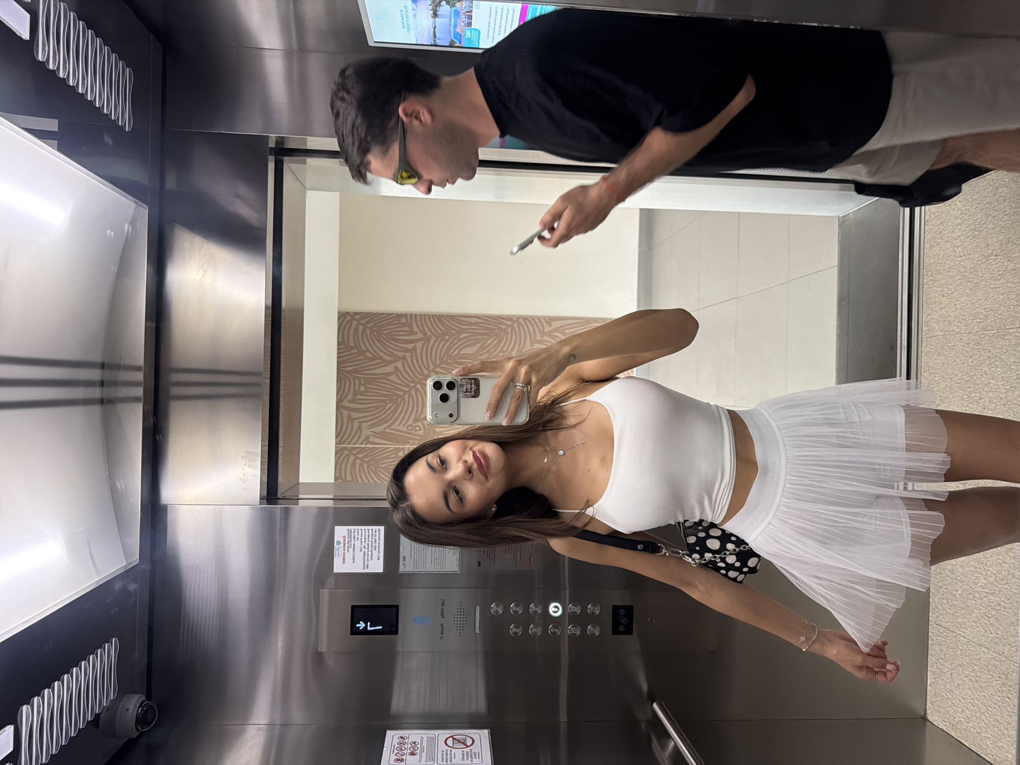 Inside an elevator, a woman with brown hair is taking a selfie with her phone. She's wearing a white top and a white tulle skirt with a black and white patterned bag. A man stands behind her, looking at his phone. He is wearing a black t-shirt and light-colored shorts. The elevator has stainless steel walls and a control panel with buttons. Outside the elevator, there's a decorated wall. An advertisement can be seen above the man. The location is likely within a building in Choeng Thale, Thailand.