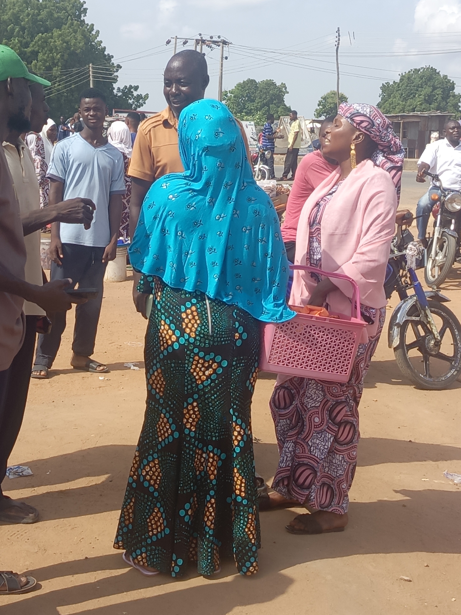 The image depicts a group of people gathered in what appears to be a marketplace in Lafia, Nigeria. The ground is dusty and light brown.
A woman in a light blue head covering and a long, dark green dress with circular orange and blue patterns is standing with her back to the camera. To her right, a woman is wearing a pink head covering, a light pink jacket, and a long pink and white patterned dress. She is carrying a pink plastic basket.
To the left, several men are standing. One is wearing a green hat and appears to be holding a phone. Behind them, other people are visible, including a man in a blue shirt.
In the background, there are trees, buildings, and power lines. A motorcycle is parked near the second woman.