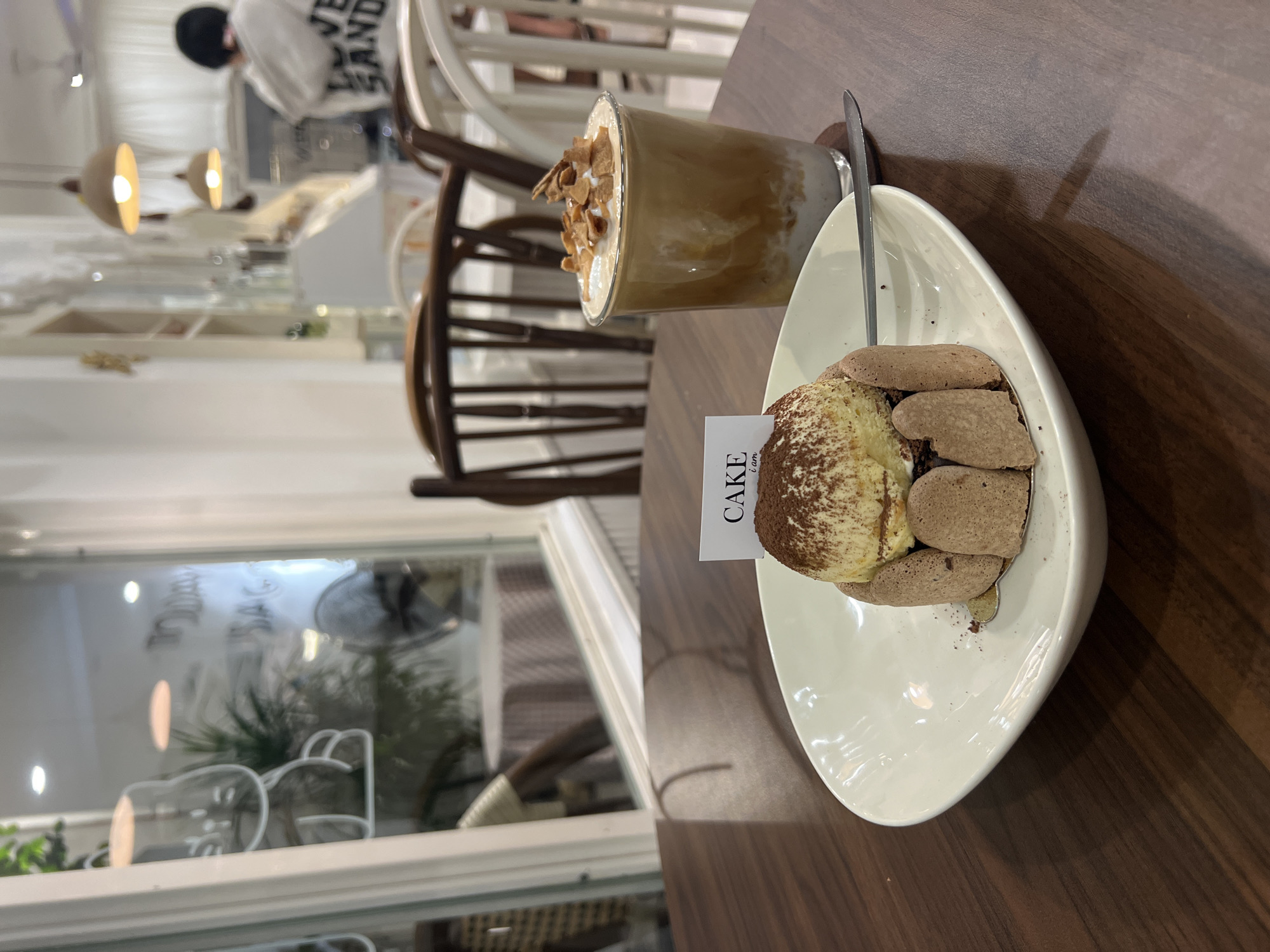 The image shows a cafe table with a tiramisu cake on an asymmetrical white plate, a drink with whipped cream and nuts, and a silver spoon. A "Cake IAM..." sign is placed in the tiramisu. The background reveals a cafe interior with chairs, a window view of plants, and a person wearing a white shirt. The scene appears to be in a cafe, likely in Nha Trang, Vietnam, based on the style of cafe interiors and common cafe items.