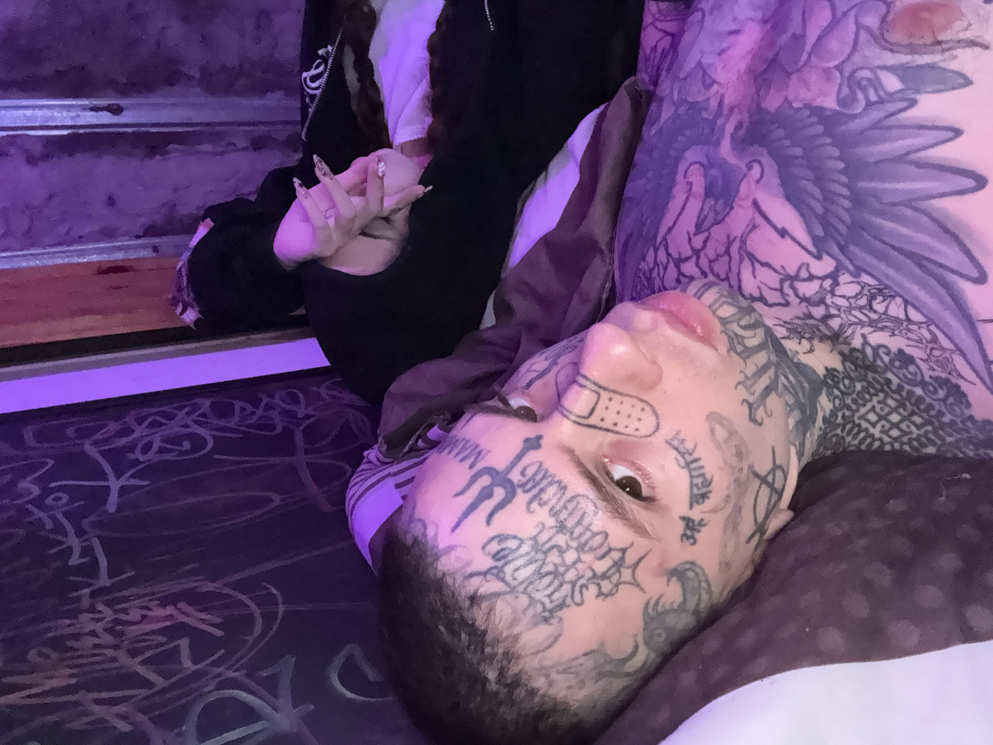 A medium shot shows a person with extensive facial tattoos lying down on a patterned surface, possibly a bed, indoors. The person's head is tilted, with their right eye visible and looking directly at the camera. A bandage is above their right eyebrow. Their body is partially visible and covered with tattoos as well, including a large tattoo on their upper chest. Another person is next to them, their hands are crossed. The background consists of what appears to be a dark, patterned surface. The lighting is dim and has a purple hue. There is no indication of a specific location in Buenos Aires, Argentina.
