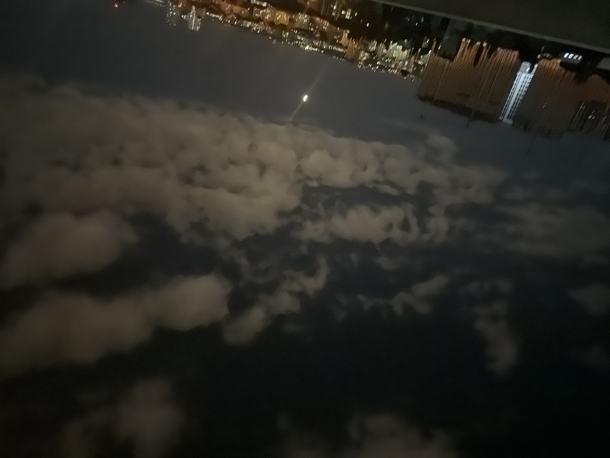 An outdoor, nighttime, high-angle shot shows a city skyline under a cloudy, dark sky with a crescent moon visible. In the foreground, a rooftop railing is present. The middle ground is dominated by buildings of varying heights, with some illuminated and others dark. The background consists of a dense urban landscape, with numerous lights indicating a populated area. The overall impression is one of a nocturnal cityscape with a prominent moon and clouds. While the image is described as being of Kyiv, Ukraine, this specific location detail is not visually verifiable from the image content itself.