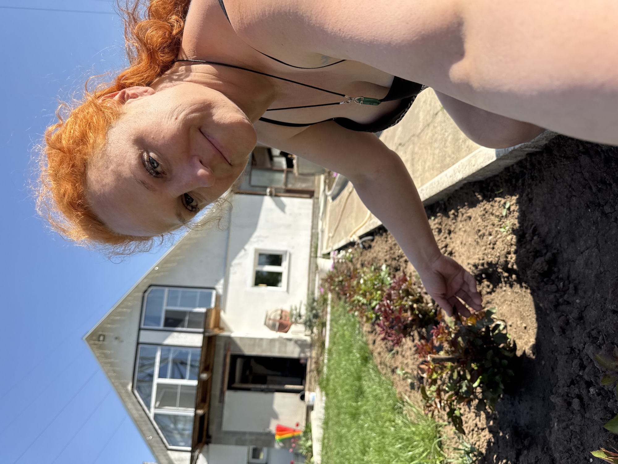 The image shows a woman with red hair, wearing a black top and a necklace with a green pendant, tending a flower bed. The flower bed contains a variety of plants with green and red leaves. Behind the flower bed, there is a white house with multiple windows, and an adjacent concrete structure. The scene is set under a clear blue sky. It is not possible to determine the specific location within the city of Chaikovsky, Russia from the given information.