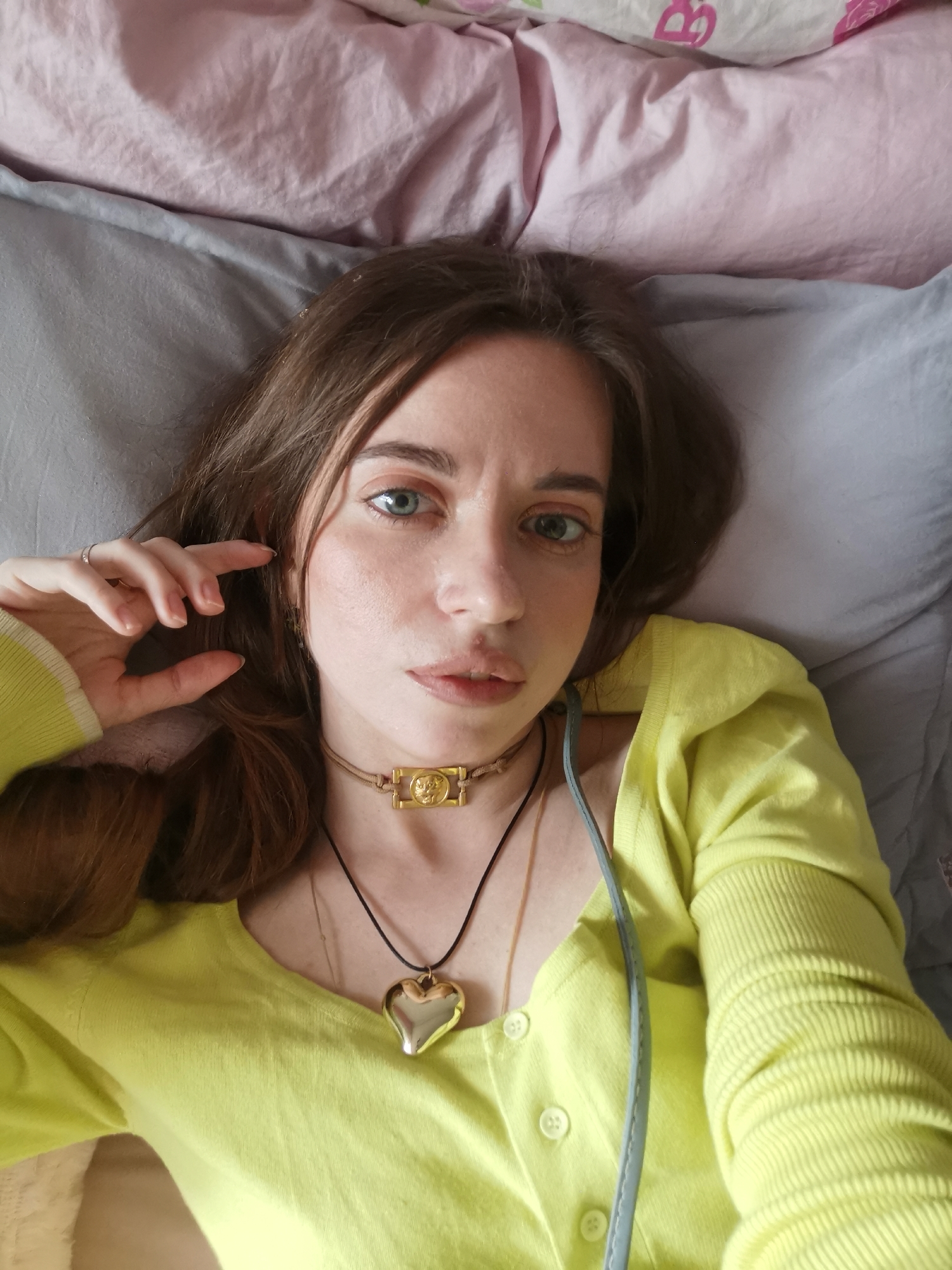 A person lies on a bed with a pink pillow on top and a gray pillow beneath them, wearing a yellow top and a choker necklace, as well as a heart necklace. The location and city are unknown.