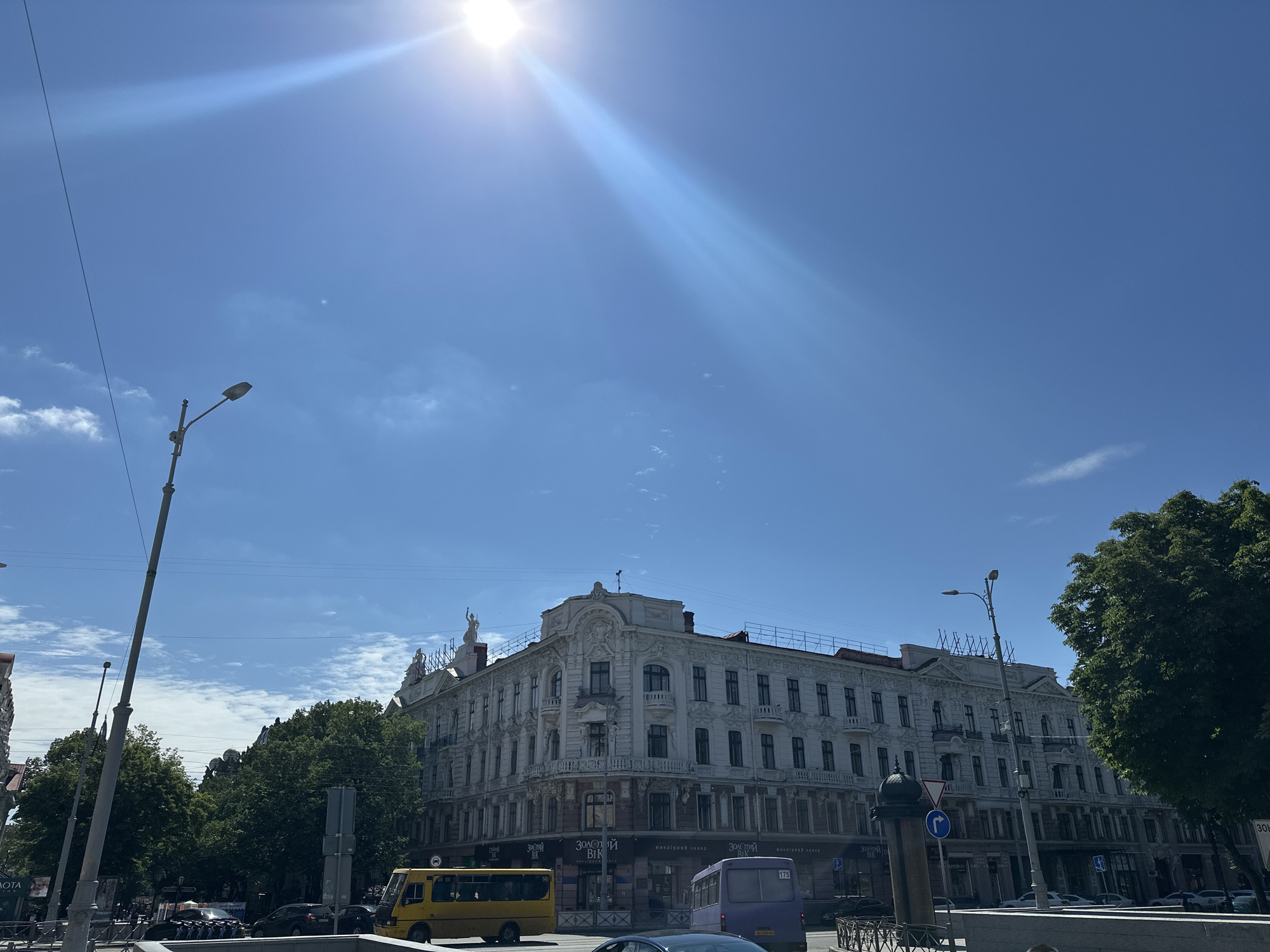 The image captures a street scene on a sunny day in an unidentified city. The primary focus is a four-story building of ornate architectural style, featuring decorative elements and multiple windows. A few buses, one yellow and one purple, can be seen on the road in front of the building. Other vehicles are also visible, suggesting a moderate level of traffic. Trees are present, adding greenery to the scene. Streetlights and utility poles are also visible in the foreground. The sky is a clear blue with some wisps of clouds, and the sun is positioned at the top of the frame, casting bright rays. The location is denoted as Unknown, Unknown.