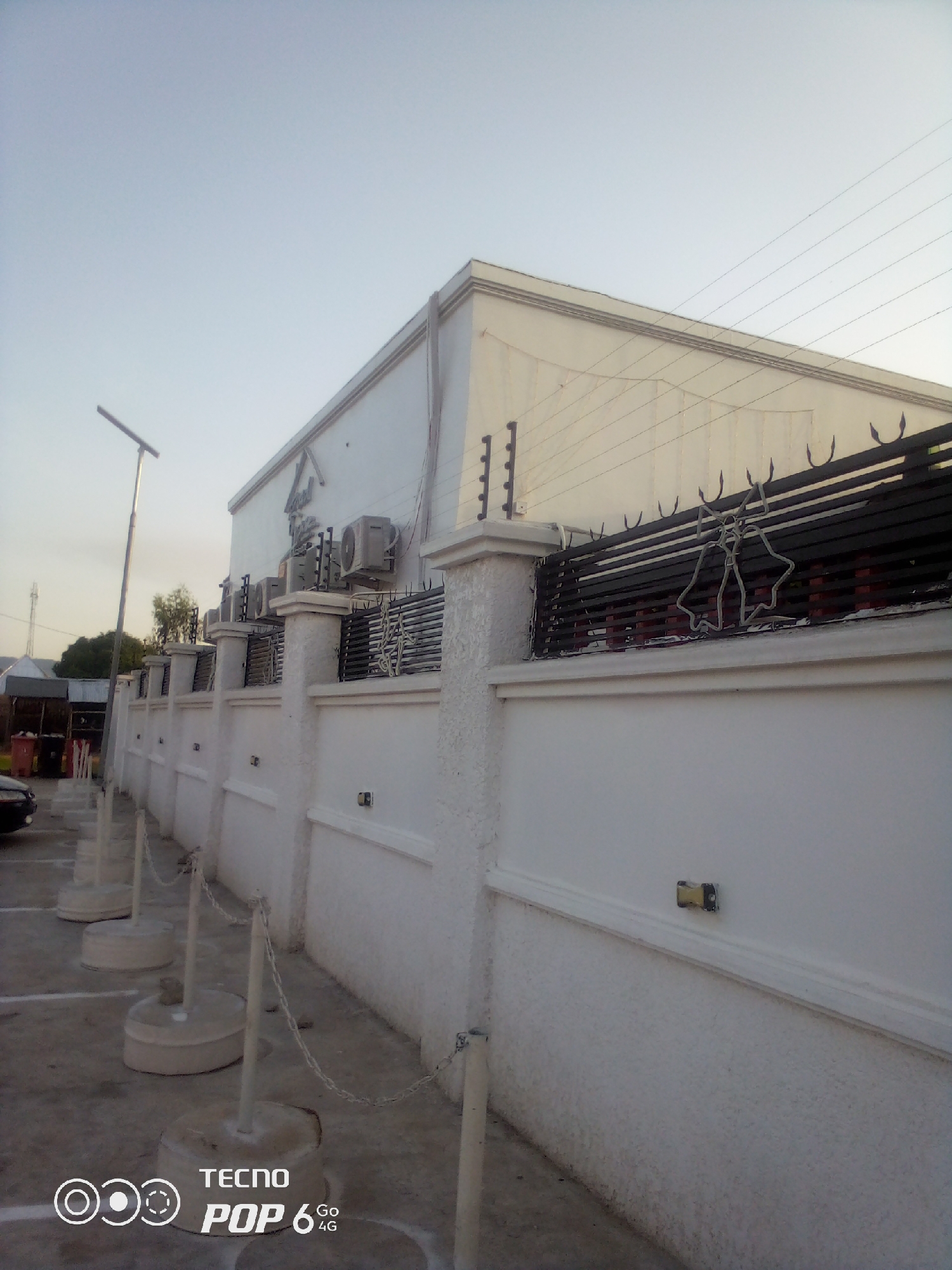 This image shows the exterior of a building with a white stucco wall and a black wrought-iron fence topped with barbed wire. Several air conditioning units are mounted on the building wall, and a street lamp is visible on the left. The fence features a decorative element shaped like two bells. In the foreground, there are several concrete bollards connected by chains, marking a parking area. The ground appears to be paved, and there are some white markings on it. A portion of a dark-colored car is visible on the far left. In the background, some trees and smaller structures can be seen. The sky is clear and light, suggesting it is either early morning or late afternoon. The scene appears to be in an urban or semi-urban setting in Howayi, Nigeria. The image also contains a watermark at the bottom left corner that reads "TECNO POP 6 4G". There is no clear activity or event taking place, and the overall mood is neutral and observational.