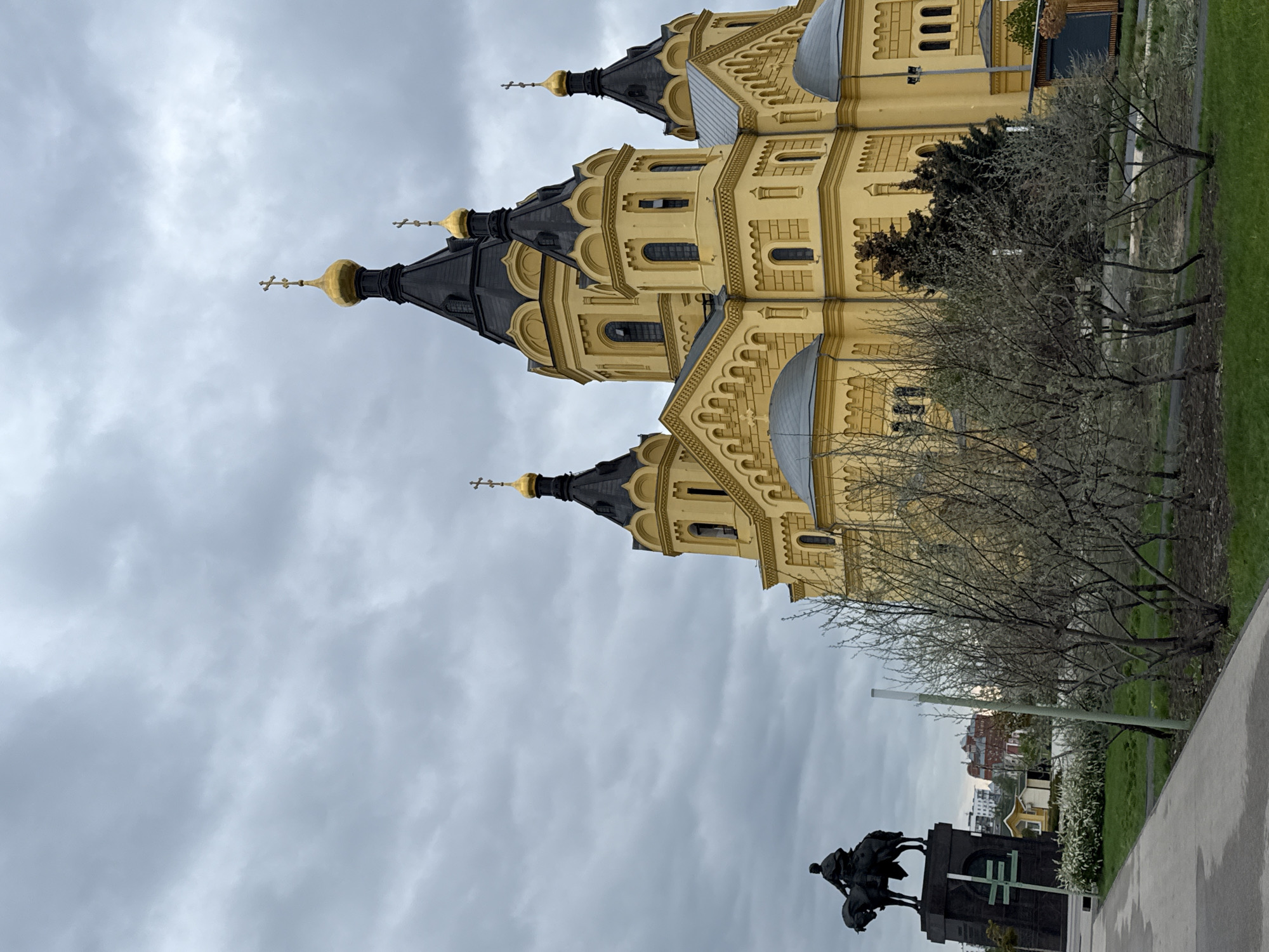 The image depicts the Alexander Nevsky Cathedral in Nizhny Novgorod, Russia. The cathedral is a large, yellow building with black domes and gold crosses on top. The sky is overcast and grey. In the foreground, there is a statue of a man on horseback. A tree stands between the statue and the cathedral. A sidewalk is in the immediate foreground, and some buildings are faintly visible in the background, behind the statue. The overall scene suggests a cloudy day in a public space with historical landmarks.