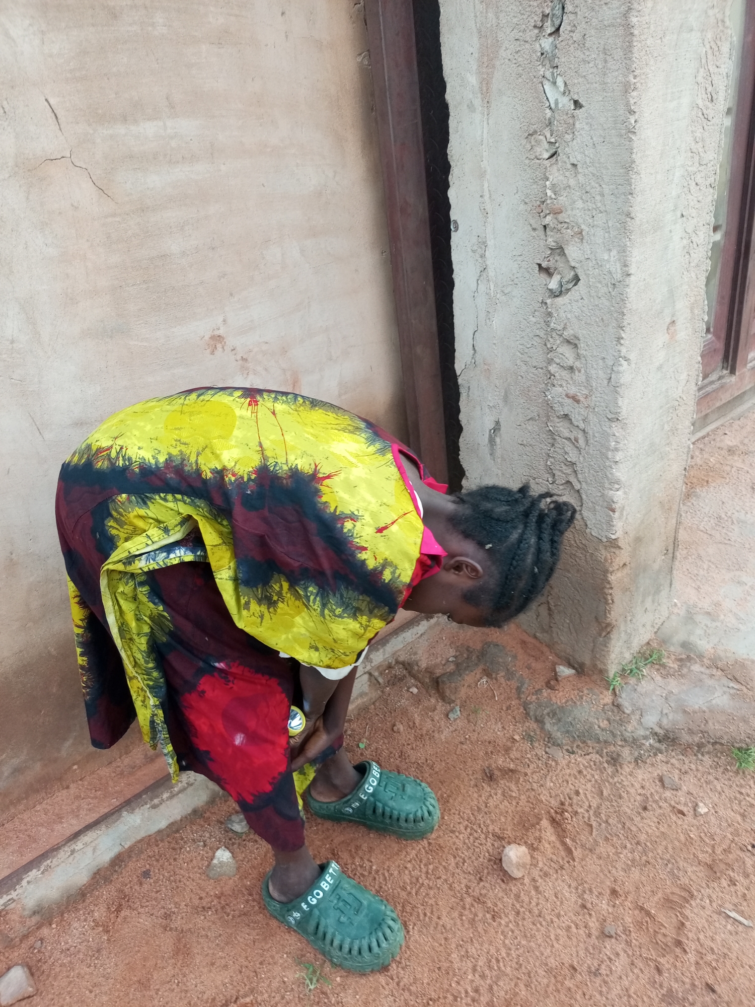 In Tumfure, Nigeria, a person is bent over, facing a textured, beige wall with visible cracks and patches of rough cement. The individual wears a brightly colored dress with a tie-dye pattern featuring yellow, red, and dark blue/black. They have dark hair braided and are wearing green clogs that have text on them. The ground is covered in reddish-brown dirt with scattered small stones and patches of green vegetation. To the right of the person, a segment of a building's foundation is visible, showing rough concrete and a wooden window frame.