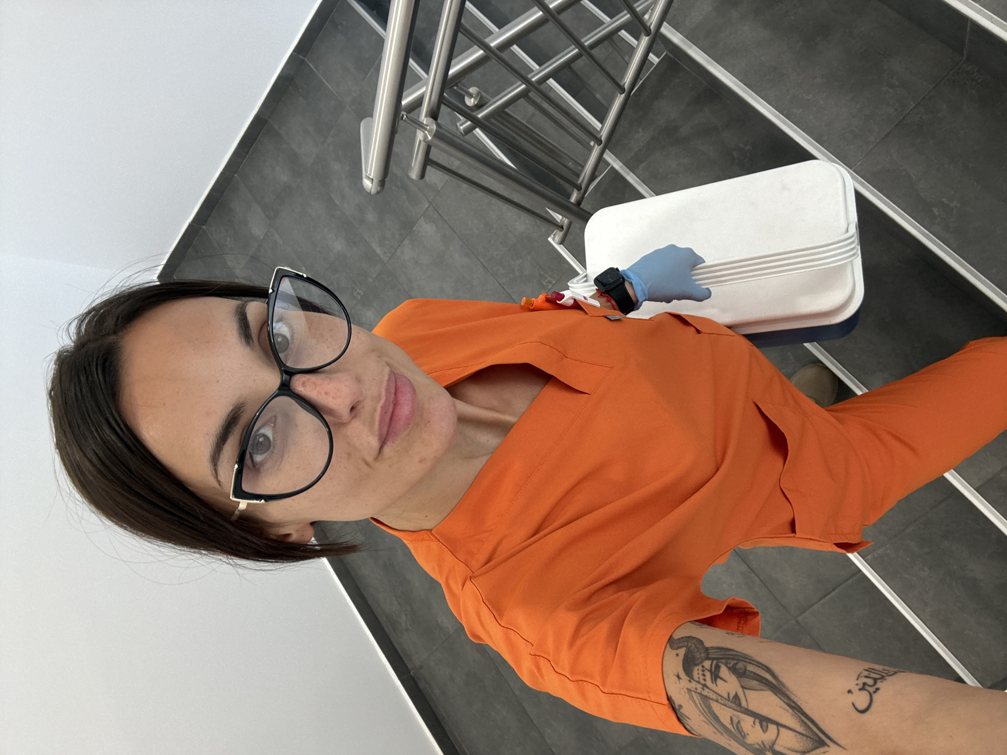 A medium, vertical, eye-level shot captures a woman indoors in what appears to be a hospital or clinic setting. The background consists of a stairwell with black and gray tiled steps and a silver handrail.
The woman, dressed in an orange medical scrub top, faces the camera, wearing large black-framed eyeglasses. She is holding a white container. Her left arm has a visible tattoo. On her right hand, she wears a blue medical glove and a black watch. Some medical items are visible in her right hand.
Based on available information, it is impossible to confirm whether this photograph was taken specifically in the city of Pleven, Bulgaria.
