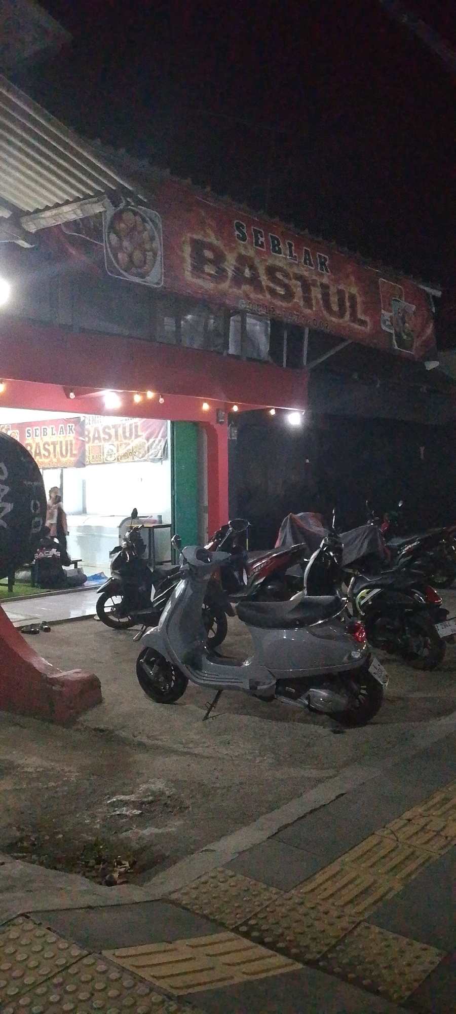At night in Banjarsari, Indonesia, a gray Vespa scooter is parked in the foreground on a paved surface. Behind it, several other motorcycles are parked, some covered with tarps. To the left, a red and white banner displays the words "SEBLAK BASTUL" and a phone number, indicating a food establishment. The building behind the banner appears to be a storefront with lights illuminating the interior. A person is visible inside, near a refrigerator or display case. The overall environment is dimly lit, suggesting it is late evening or night. The pavement in front of the building features tactile paving for the visually impaired.