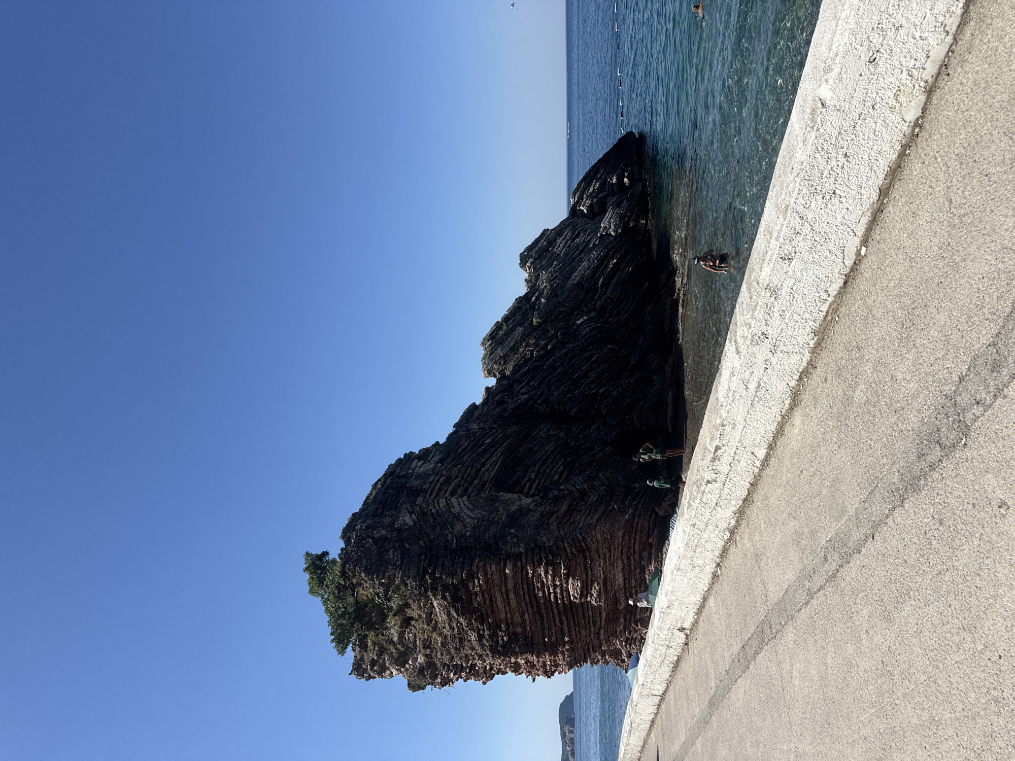 In Rafailovići, Montenegro, a large layered rock formation juts out from the sea near a paved walkway lined with a white stone barrier. The sky is a clear, vibrant blue. Several people are visible in and around the water. Two individuals are standing near the base of the rock, while others are swimming. One person is in the shallow water near the shore with a snorkel mask. The sea is calm and appears clear, with a roped-off swimming area marked by buoys in the distance. The walkway provides a view of the rock and the sea.