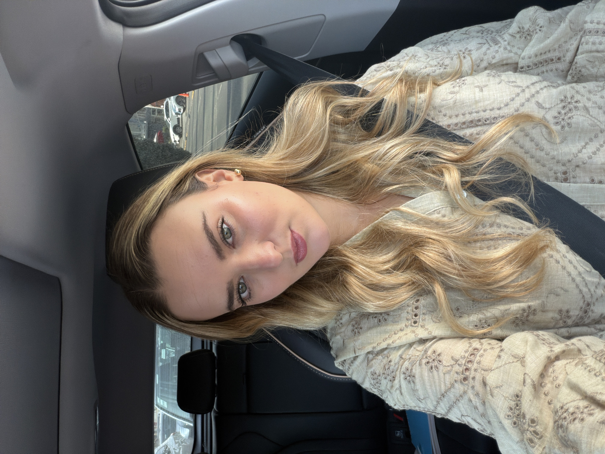 A fair-skinned woman with long blonde hair is sitting in a car. She is wearing a patterned cream-colored shirt and a seatbelt. The car is driving down a street in Puebla, Mexico. Buildings are visible in the background.