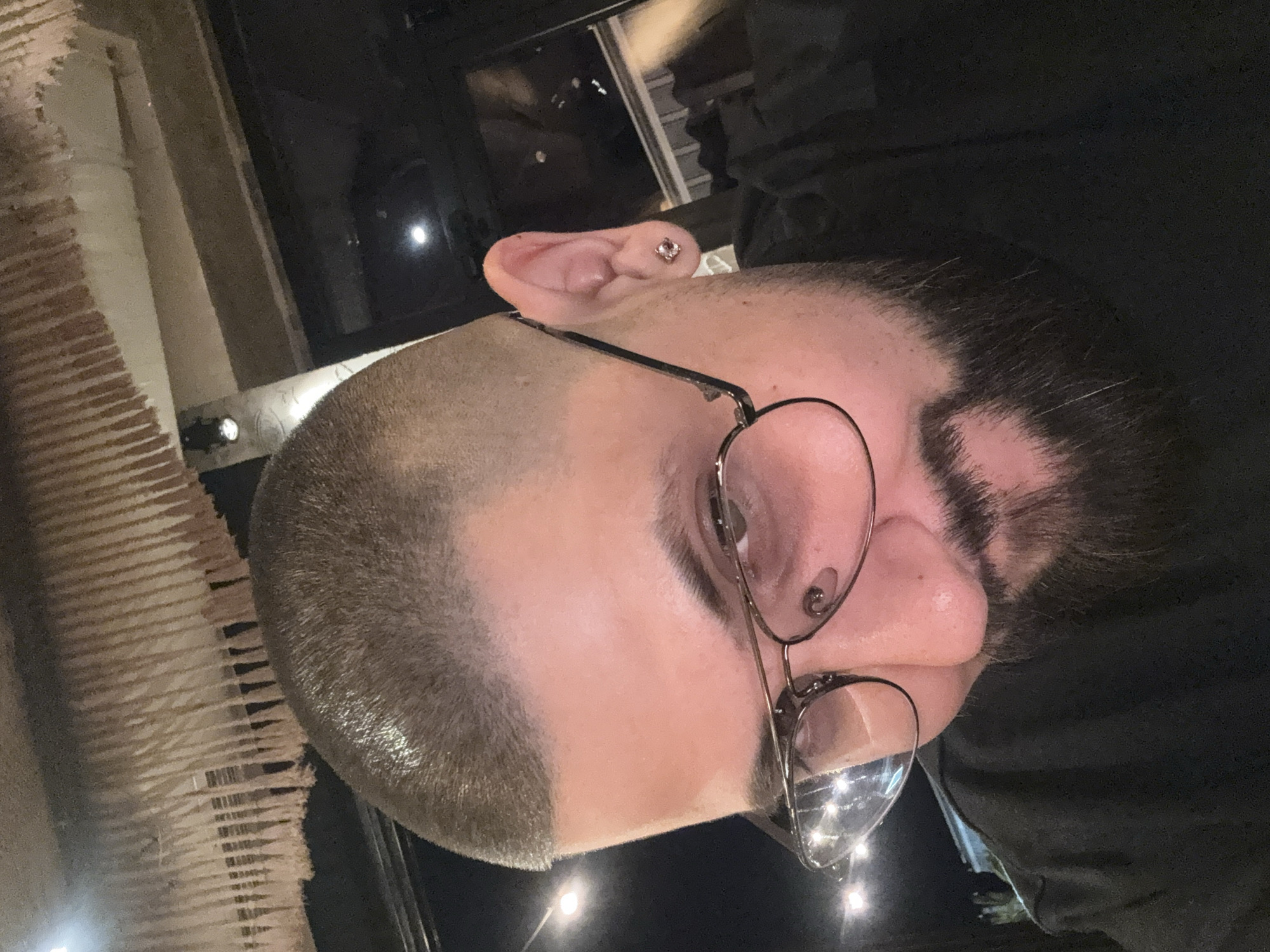 The image is a close-up selfie of a man with a buzz cut and a beard. He is wearing glasses with thin metal frames and a black t-shirt. He has a small stud earring in his right ear. The background appears to be indoors, with some decorative elements hanging from the ceiling, possibly tassels. There are also lights visible in the background. The man is looking down and to his left, with a slight smile. The image does not contain any specific geographical indicators that would allow for its localization to Varna, Bulgaria, or any other city.