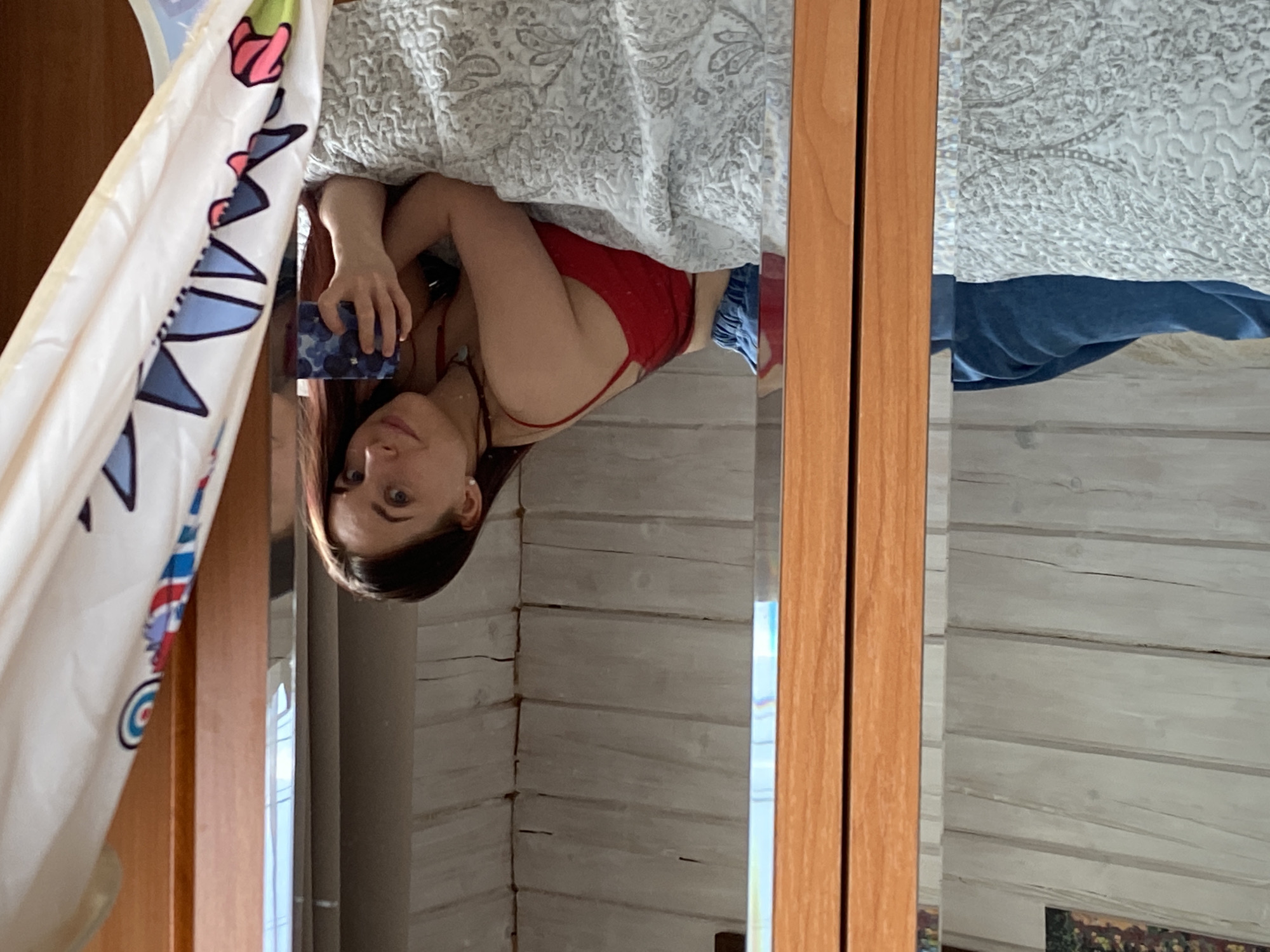 The image shows a woman reflected in a mirror. She is partially under a bed, wearing a red top and blue shorts. She holds a phone in her hand, presumably taking the picture. The background appears to be a room with log walls.