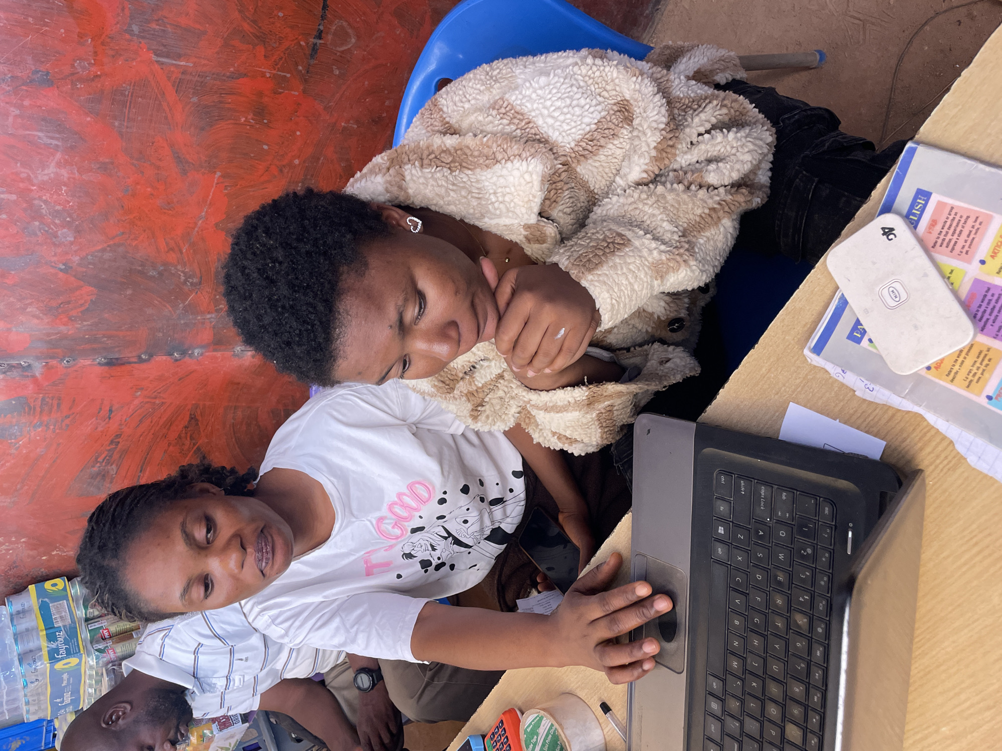Two African women are positioned at a wooden desk, with one seated on a blue plastic chair. The woman on the left is actively using a partially open laptop. Various items such as water bottles, notebooks, and office supplies are scattered on and around the desk. A white phone and a 4G device sit on a notebook, with English notes visible on the pages. An older African man is also visible in the lower left corner, with a partial view of another person behind the woman using the laptop. The setting is Gura Top, Nigeria.