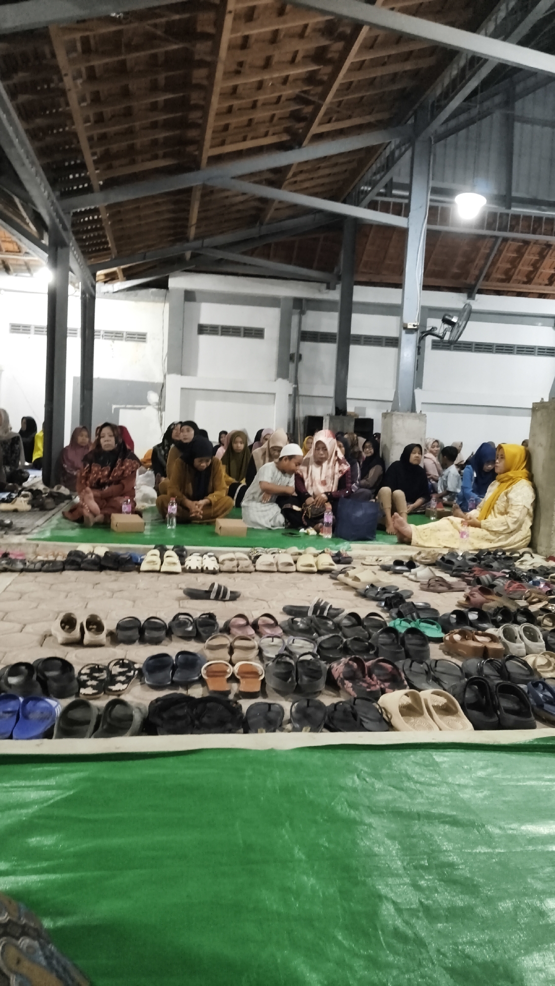 The image depicts a gathering of people, predominantly women and a few children, seated on a green tarp or mat inside a building with an exposed wooden roof structure. The environment appears to be an indoor space, possibly a community hall or a religious gathering place, given the attire of the individuals and the presence of shoes neatly arranged in rows in the foreground and on the ground. The people are dressed in traditional Islamic clothing, with many wearing hijabs. Some individuals are engaged in conversation, while others appear to be listening or observing. The lighting in the scene is somewhat dim, with artificial lights visible in the upper portion of the image. Rows of footwear, including sandals and flip-flops, are arranged on the floor, suggesting that participants have removed their shoes before entering the area. The overall impression is of a communal gathering, possibly for a religious service or a community event, taking place in Dalegan, Indonesia.