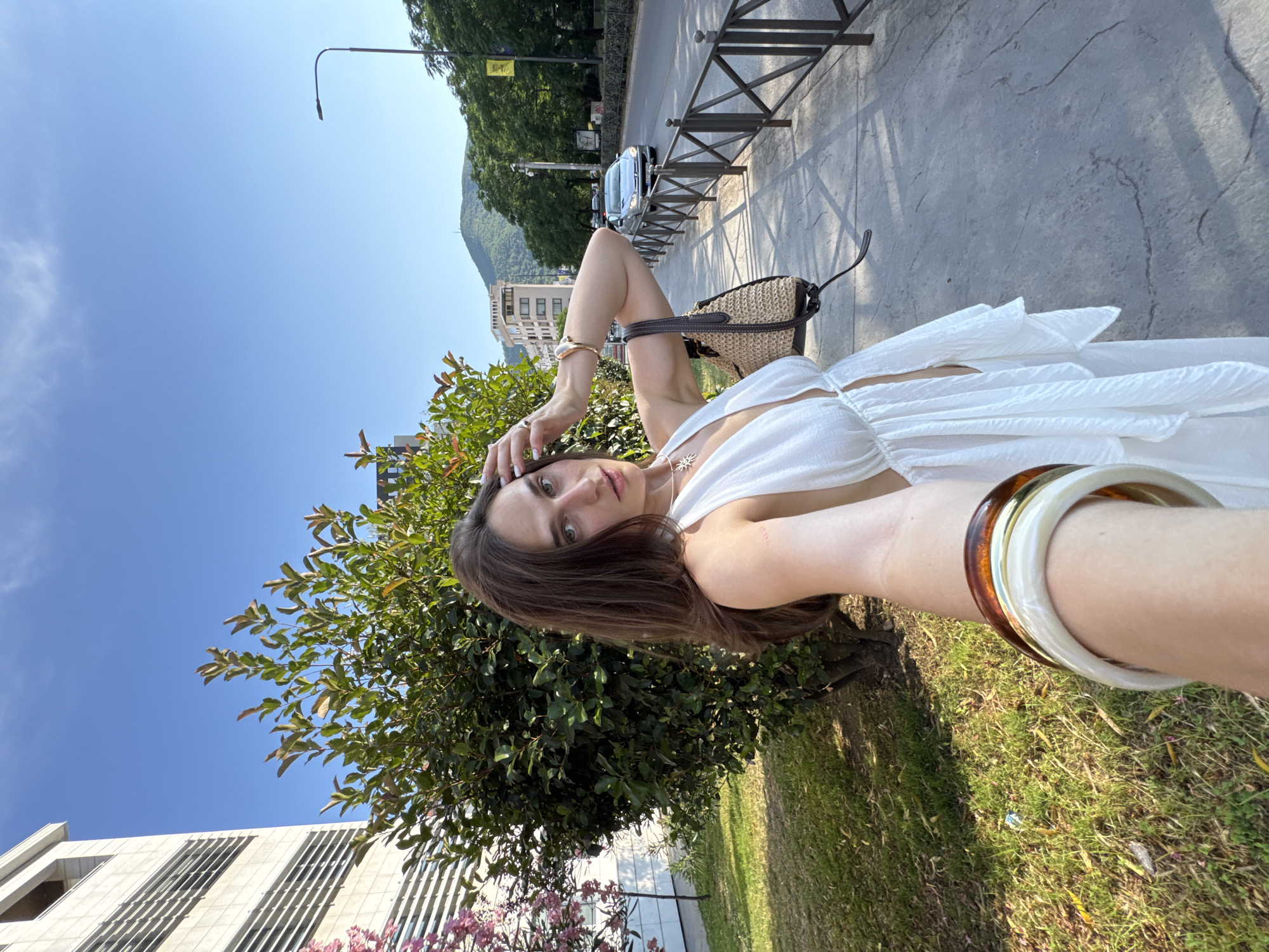 This media file captures a young woman taking a selfie outdoors on a sunny day, likely in Budva, Montenegro. She is wearing a white dress and has her hair down. She is accessorized with bracelets on her arm and a necklace. A straw bag is visible behind her. The background includes trees, a building, a street with a car, and a body of water in the distance, suggesting an urban or semi-urban environment near the coast. The sky is clear and blue, indicating good weather. The lighting suggests it is daytime, possibly late afternoon given the shadows. The woman's expression is bright and engaging, conveying a sense of happiness or confidence. There are no visible text elements in the image.