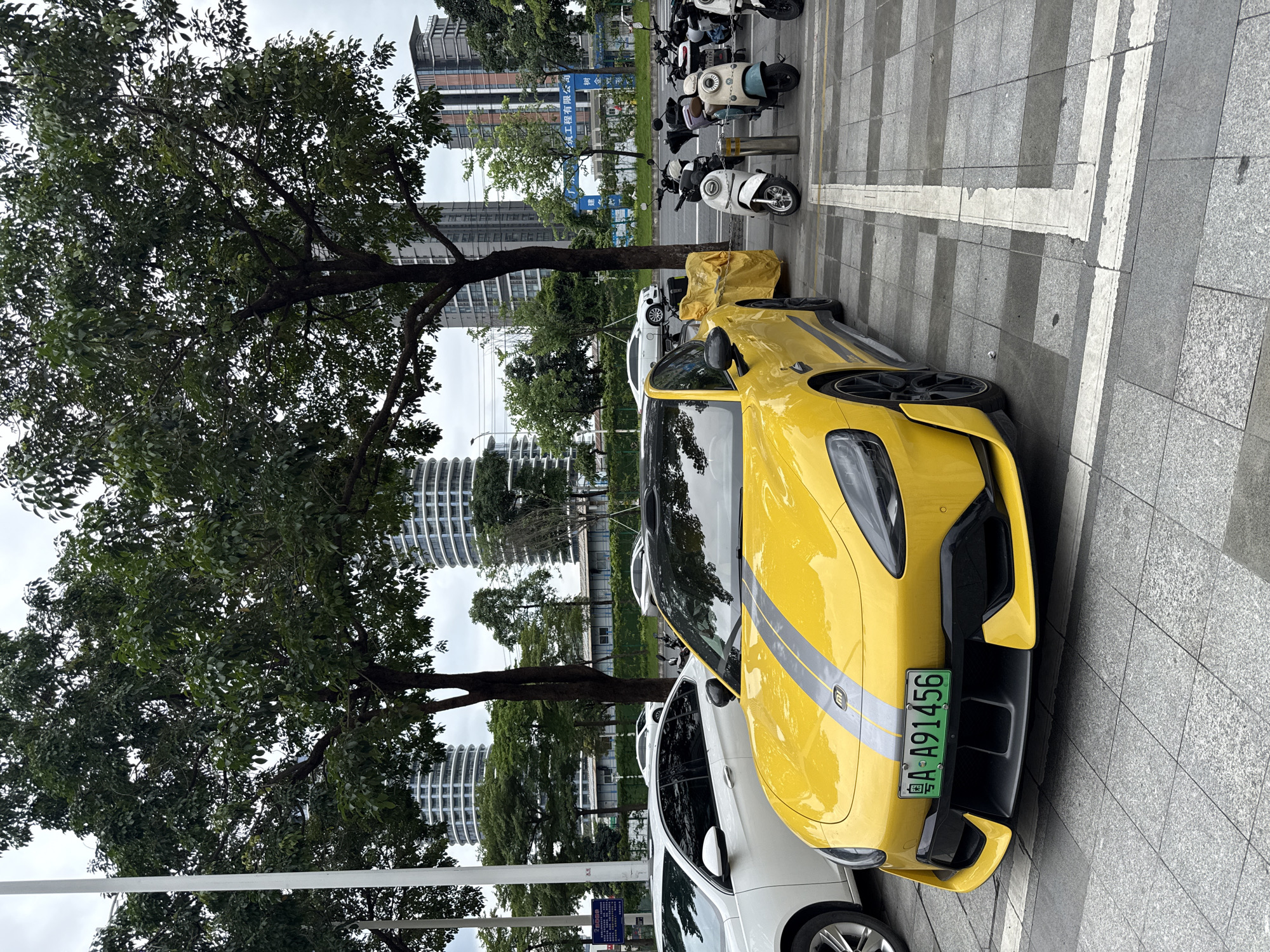 A bright yellow sports car with grey racing stripes is parked on a paved lot in Tianhe District, China. The car has a black roof and black accents on the front bumper and side skirts. Its license plate reads "A A91456". To the left of the sports car is a white sedan. Behind the cars, several motorcycles and scooters are parked, and beyond them are tall, modern buildings and trees. The sky is overcast.