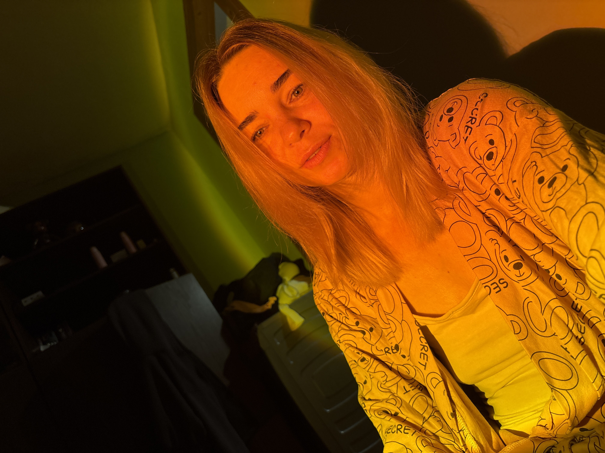 A young woman with long, blonde hair is captured in a selfie-style photograph. She is wearing a yellow top with a repeating pattern of cartoon bears and the word "SECRET" printed on it, along with a plain yellow undershirt. Her expression is neutral, and she is looking directly at the camera. The lighting in the image is warm and orange-hued, casting shadows and creating a dramatic effect. The background is dark, with hints of a room and possibly some furniture. The image does not provide any information that would indicate the location as Sinyavino, Russia.