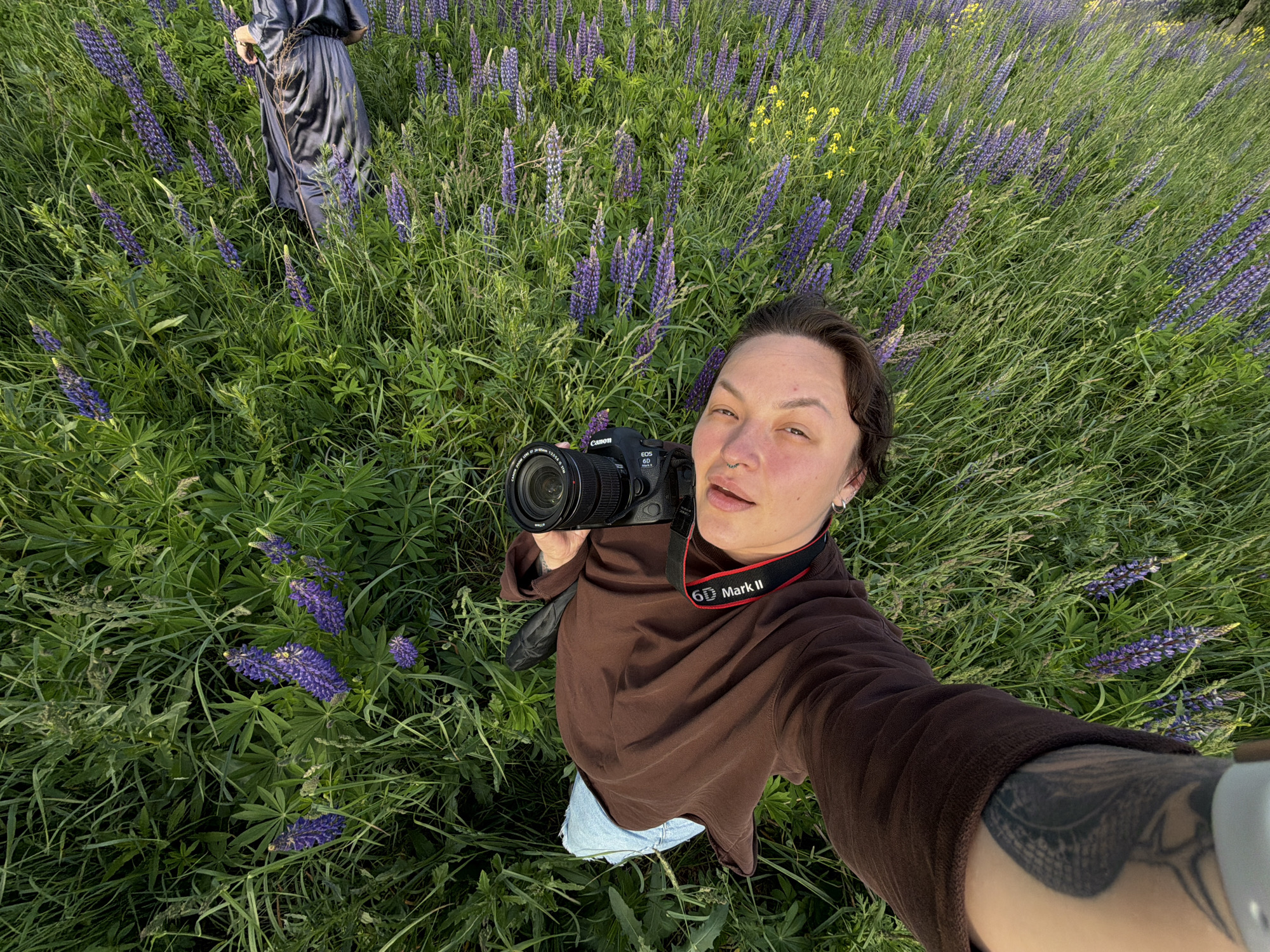 A person is lying on their back in a field of tall green grass and purple lupine flowers. They are holding a black Canon DSLR camera in their right hand, pointing it towards themselves for a selfie. The person is wearing a brown long-sleeved shirt and light blue shorts. A tattoo is visible on their left arm. In the upper right corner, a portion of a dark grey or black fabric, possibly a skirt or dress, is visible, suggesting another person may be present. The scene appears to be outdoors in a natural environment. The location is identified as a field in the city of Bolshaya Usa, Russia.