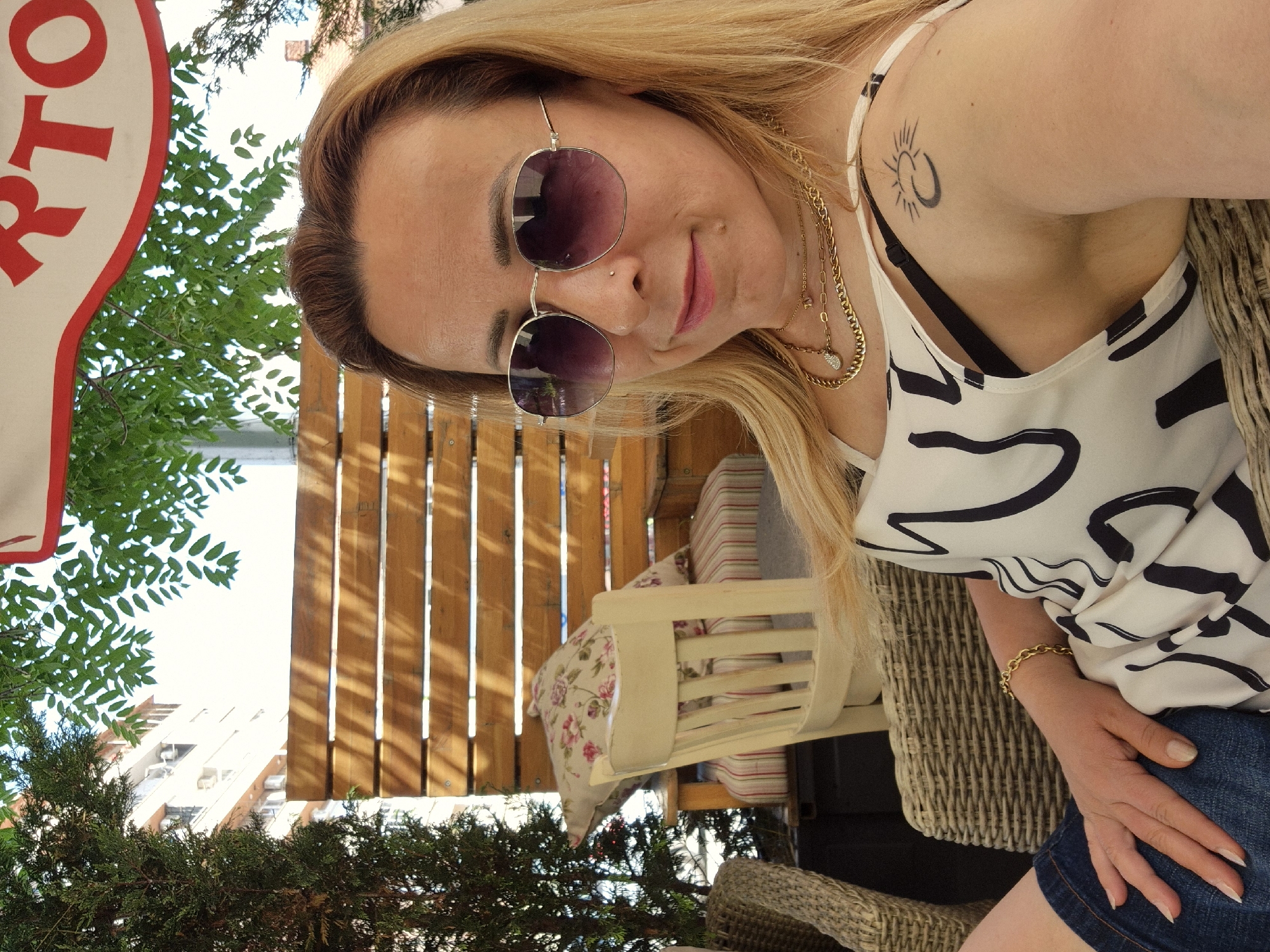 A fair-skinned woman with blonde hair and sunglasses is taking a selfie outdoors. She is wearing a white tank top with black abstract patterns, a black bra, denim shorts, and gold necklaces. She has a tattoo of a crescent moon with a sun on her right shoulder. The woman is sitting in a wicker chair with her arm resting on her lap. Behind her, there is a wooden slatted wall, a chair with striped cushions and a floral pillow, and greenery. A portion of a red and white awning with the letters "RTO" is visible at the top of the frame. The scene appears to be an outdoor seating area, possibly at a cafe or restaurant.