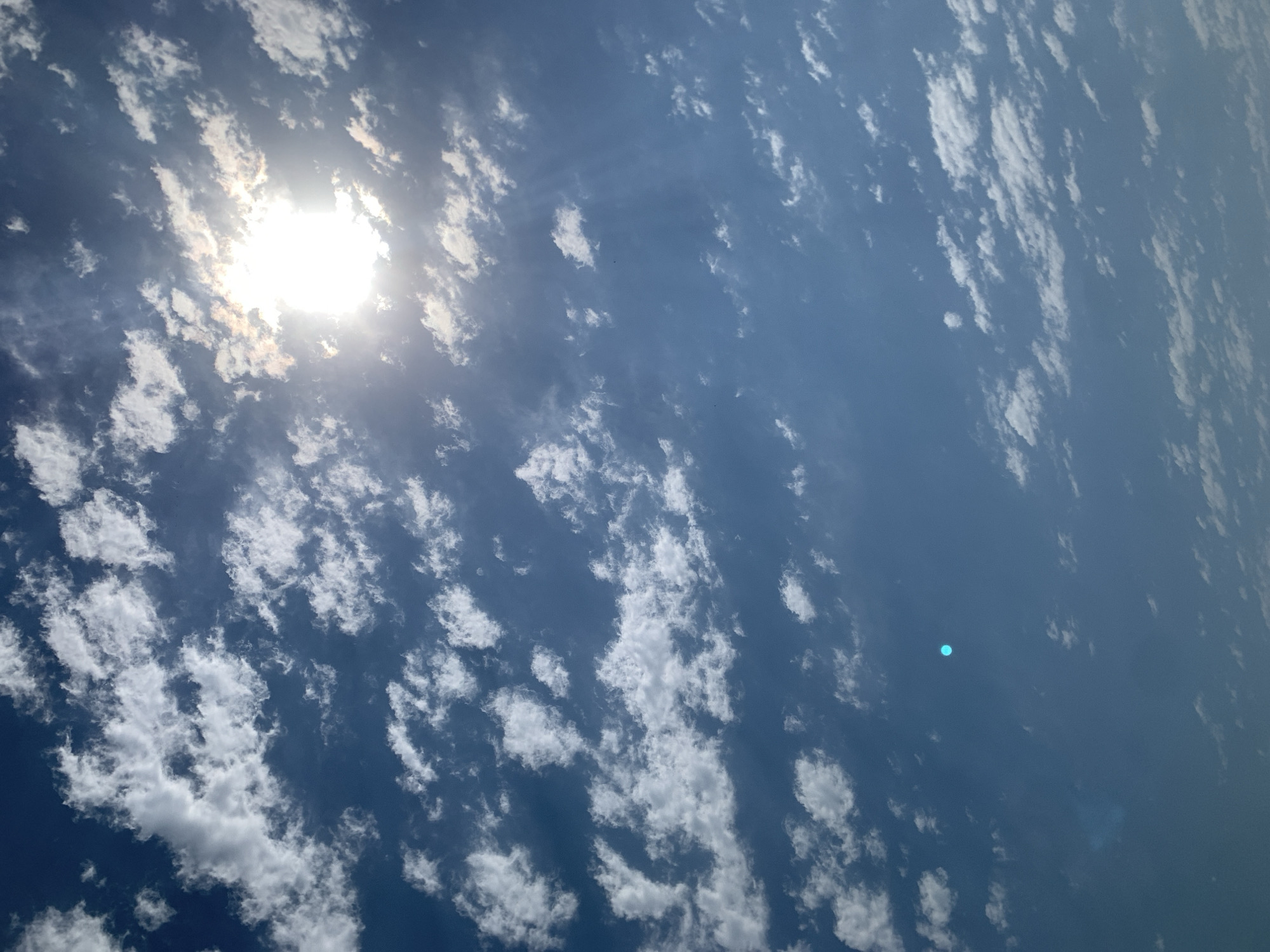 A bright sun shines through a blue sky filled with scattered white clouds. There is a small, bright turquoise circular object in the lower left quadrant of the image. The scene appears to be outdoors in Khazar Raion, Azerbaijan.