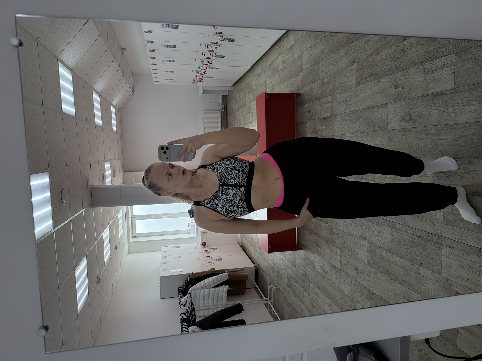 A woman in athletic attire is taking a selfie in a mirror within a room. She is wearing a patterned sports bra, black sweatpants with a visible pink waistband, and white socks. The room features lockers along one wall, some decorated with stickers, and what appears to be a clothing rack with items hanging on it. There is also a red bench. The lighting is bright, possibly from fluorescent fixtures in the ceiling. The reflection suggests the setting is a changing room or locker area, possibly in a gym or sports facility. The city of Izhevsk, Russia is not discernible from the image.