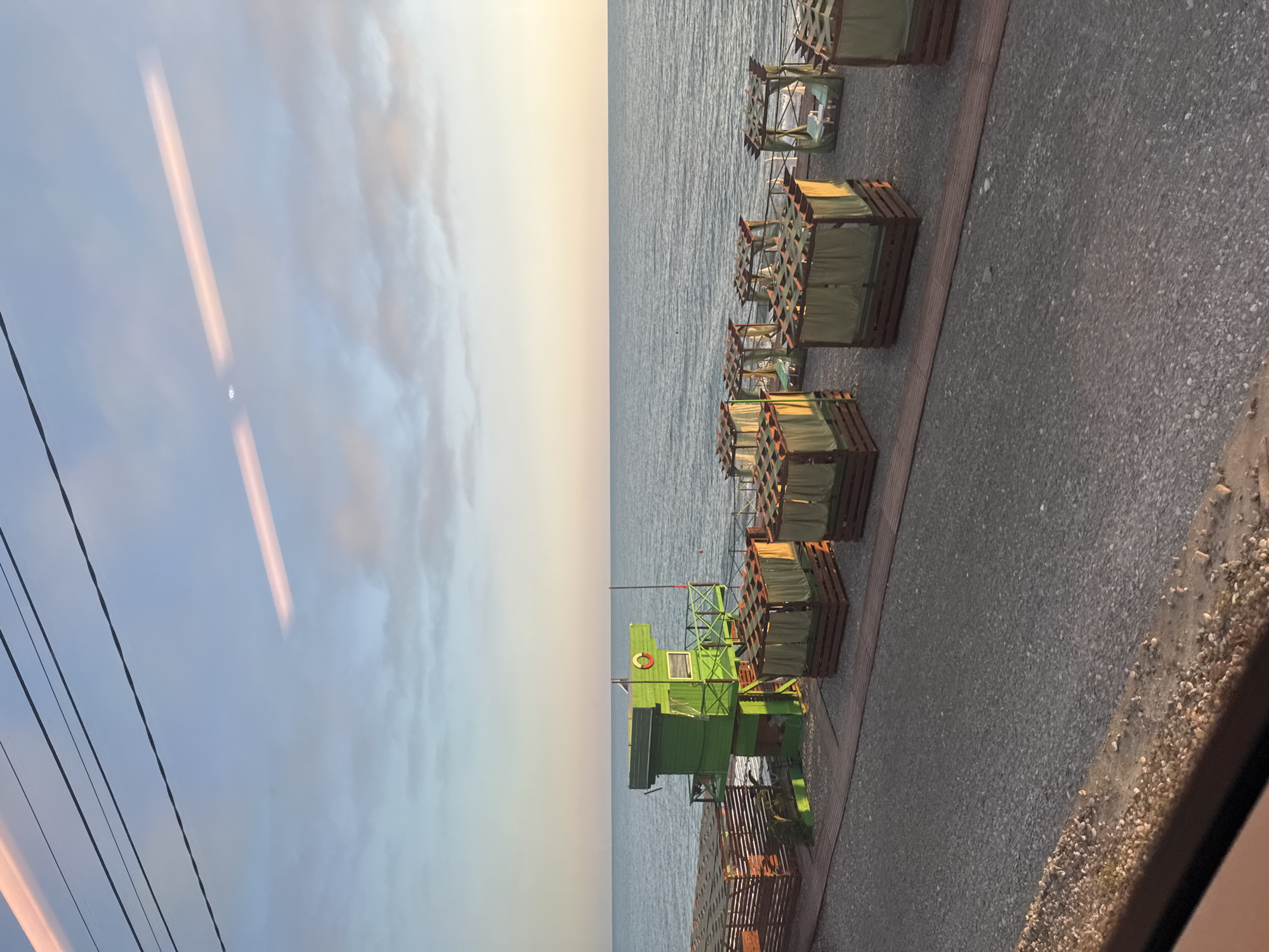 This media file depicts a serene seaside scene at what appears to be either sunrise or sunset, given the soft, diffused light coloring the sky and water. The setting is a beach or promenade area, characterized by a gravelly ground surface and a wooden boardwalk bordering the water.
Dominating the right side of the frame are rows of wooden beach cabanas, each with green fabric coverings, suggesting a resort or recreational area. These structures are arranged neatly, indicating an organized setup for beachgoers. In the foreground, a vibrant green lifeguard station stands prominently, equipped with a window and a lifebuoy, reinforcing the beach setting and hinting at safety provisions. The station has a rustic, perhaps slightly weathered appearance.
The sea stretches out in the background, a vast expanse of calm water reflecting the gentle sky. The sky itself is a gradient of soft blues and pale yellows, with some wispy clouds adding texture. The reflection of what looks like electric cables or wires is visible in the upper left portion of the image, superimposed over the sky, suggesting the viewpoint is from a window or through a reflective surface.
There is no visible text in the image. The overall mood is tranquil and peaceful, evoking a sense of calm and relaxation associated with a quiet day by the sea. The time of day is likely early morning or late evening due to the quality of light. The presence of beach amenities like cabanas and a lifeguard station points to a coastal location, potentially a tourist destination.