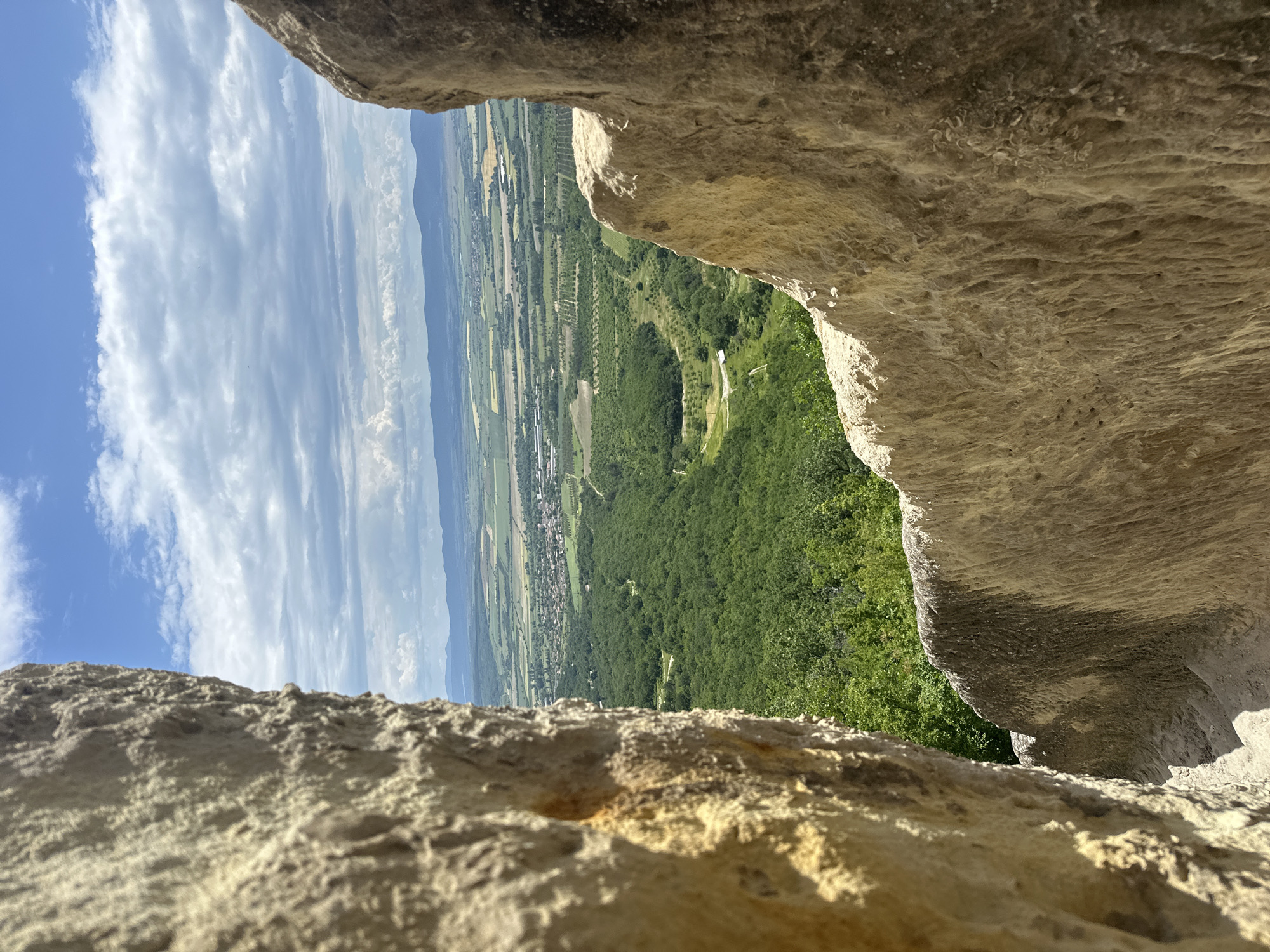 The image is taken from a rocky vantage point in Troitsa, Bulgaria, looking out over a landscape featuring a mix of forests, agricultural fields, and a small town. The foreground consists of rough, light-colored rocks, likely part of a cliff or rock formation. The view extends to a wide valley with cultivated lands showing a grid pattern. Patches of dense green forest are interspersed within the valley. In the distance, a line of low mountains can be seen under a sky with scattered clouds and blue sky. The perspective suggests the camera is positioned behind or within a rock opening, framing the landscape beyond. The image is taken during daylight hours with clear visibility.